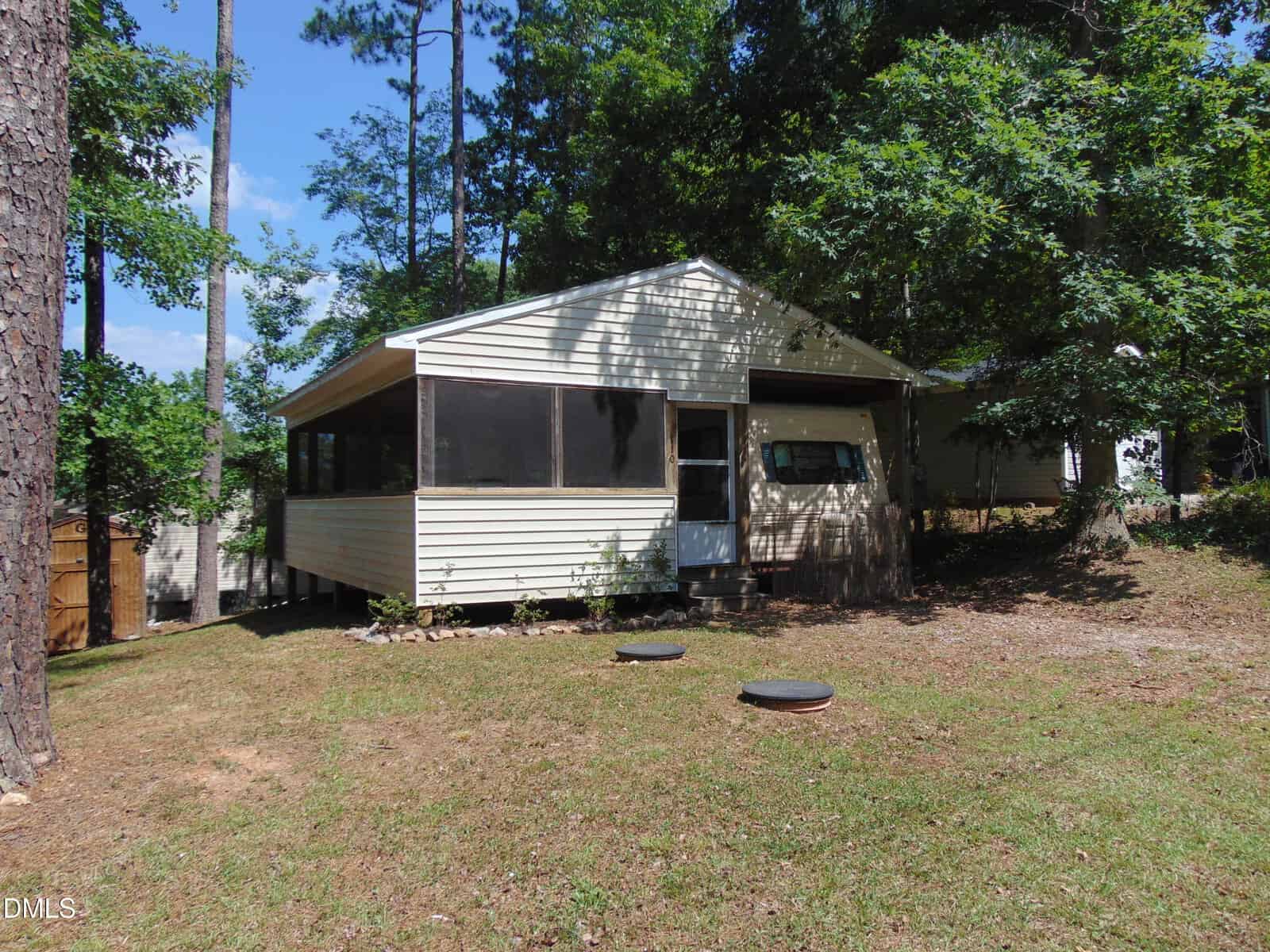 A small beige house at 110 Cimarron Drive, Louisburg, features a screened porch nestled among tall trees and grass on a slightly sloped lot. The partially shaded yard includes two round septic lids visible in front of the house.
