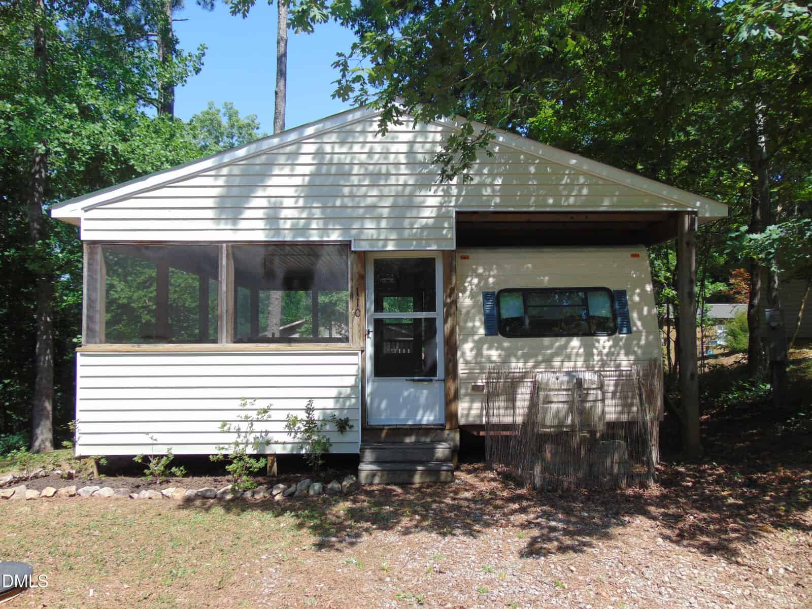 A small, single-story house with white siding, a screened porch on the left, and a metal awning over part of the front. Surrounded by trees and greenery, with sunlight casting shadows on the ground.