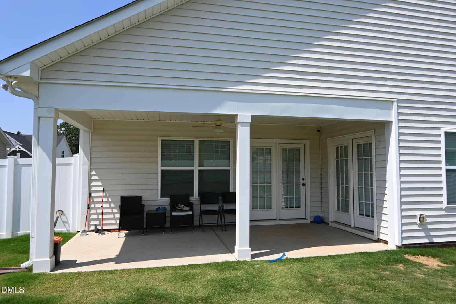 A covered backyard patio with white siding, two support columns, patio furniture, a ceiling fan, and several glass doors, opening onto a grassy lawn with a white fence along the property line.