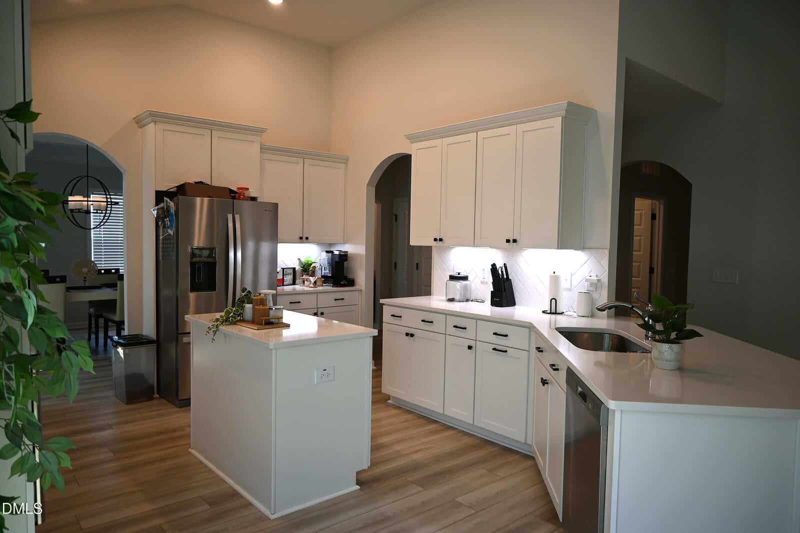Modern kitchen with white cabinets, stainless steel appliances, an island with a plant, and light wood flooring. Under-cabinet lighting brightens the counters. There's a dining area visible through an arched doorway on the left.