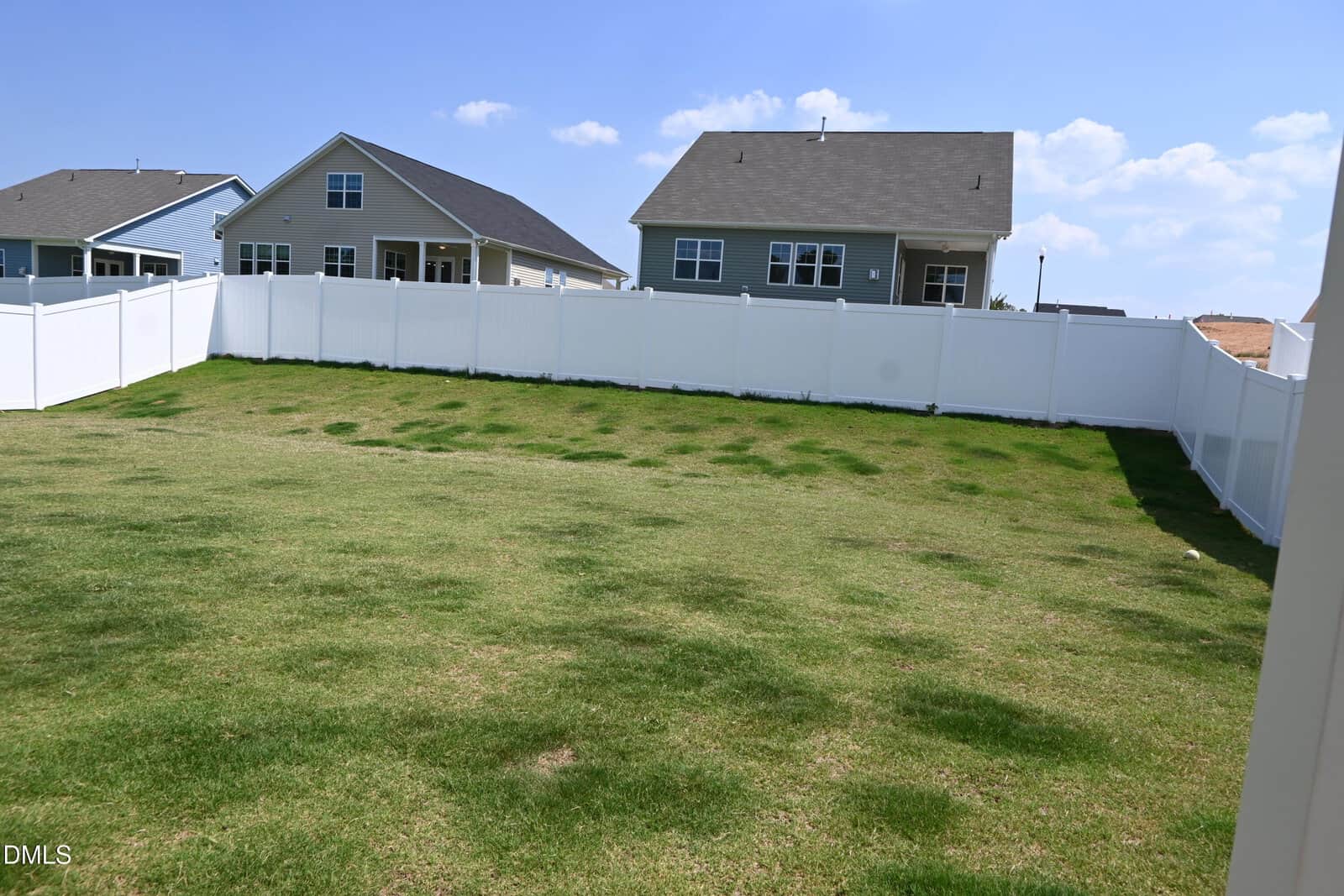 A fenced backyard with patchy grass, bordered by a white vinyl fence. Two modern suburban houses with gable roofs are visible in the background under a blue sky with some clouds.