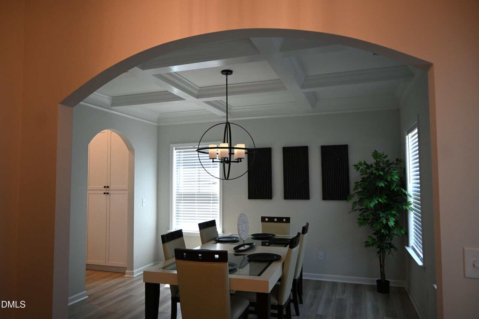 Modern dining room with a rectangular table set for six, a black chandelier, three black wall art pieces, a tall potted plant, light wood flooring, and two large windows. An arched doorway leads to a white cabinet area.