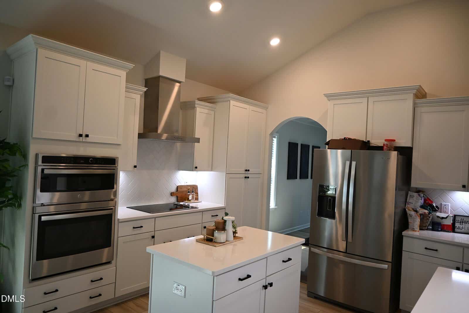 Modern kitchen with white cabinets, stainless steel appliances, an island in the center, and a range hood above the stove. Refrigerator and double oven are on the right and left. Countertops are uncluttered, and lighting is bright.