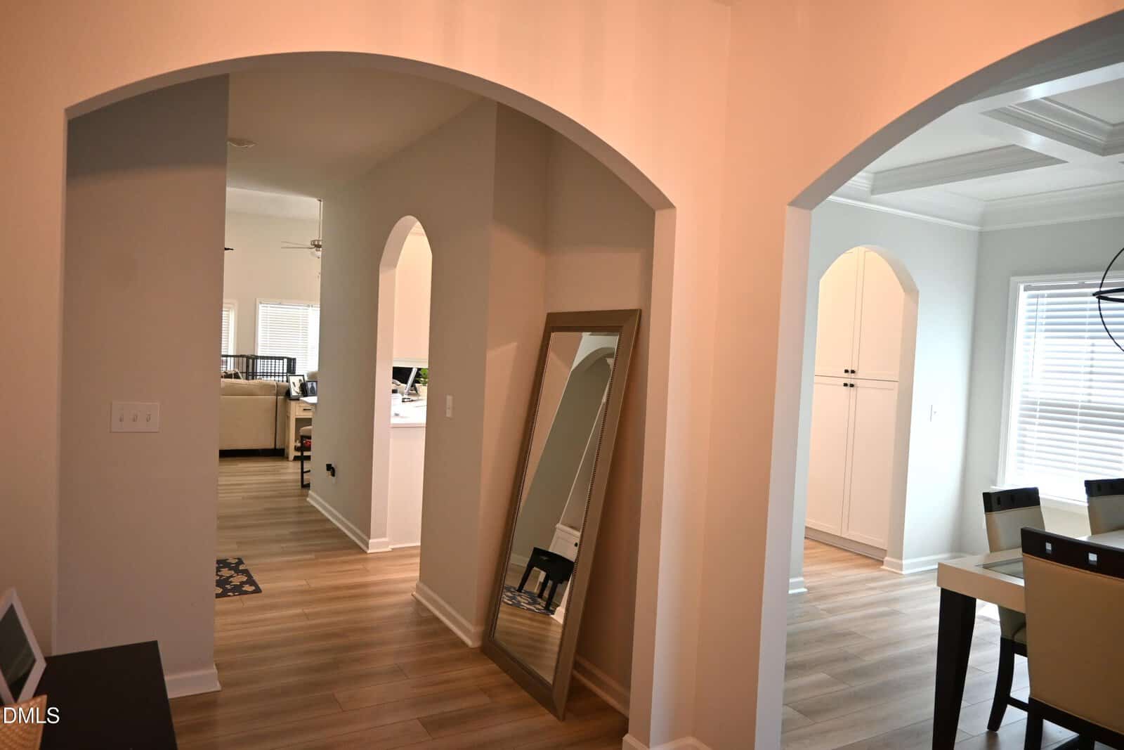 A hallway with light wood floors and arched doorways leads to a living room and dining area. A large floor mirror leans against the wall, and natural light fills the space from several windows.