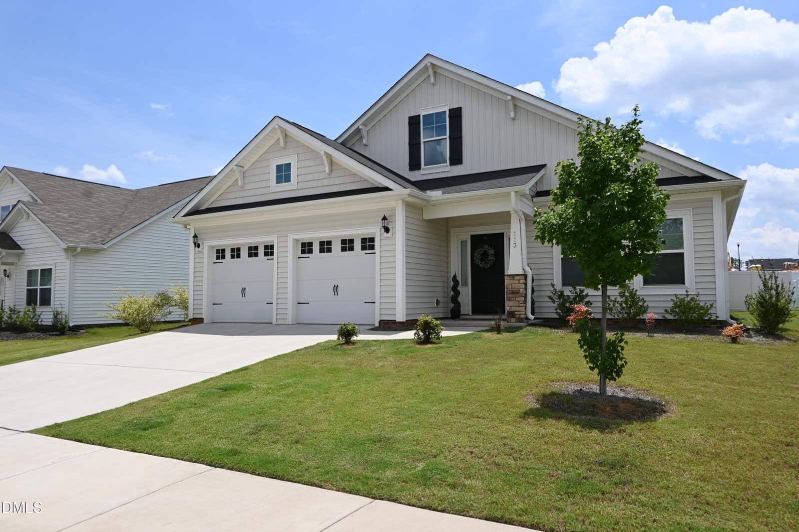 A modern two-story house with white siding, black shutters, a double garage, and a manicured lawn with young trees and shrubs under a partly cloudy blue sky.