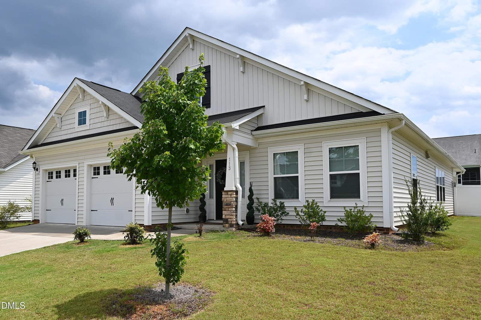 A single-story white house with a double garage, light gray roof, small front porch, and neatly landscaped yard with a young tree and shrubs, under a partly cloudy sky.