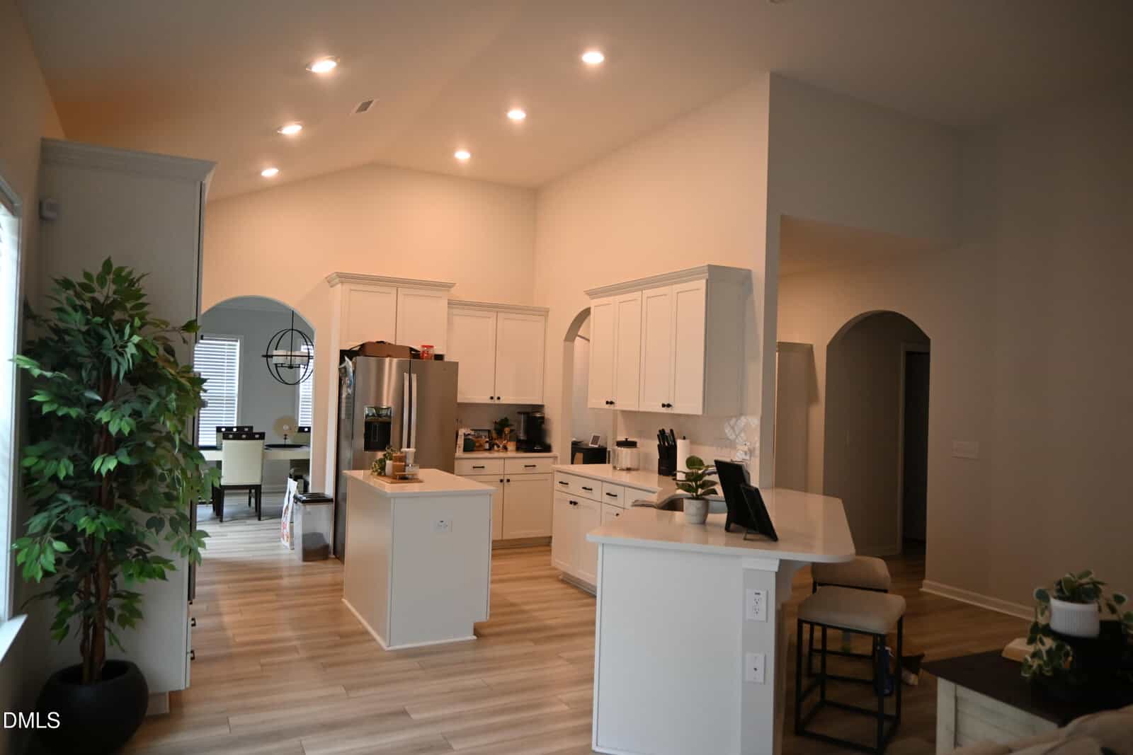 Modern kitchen with white cabinets, stainless steel appliances, an island, and light wood floors. Dining area visible in the background; potted plant and bench seating on the side. Warm recessed lighting brightens the space.