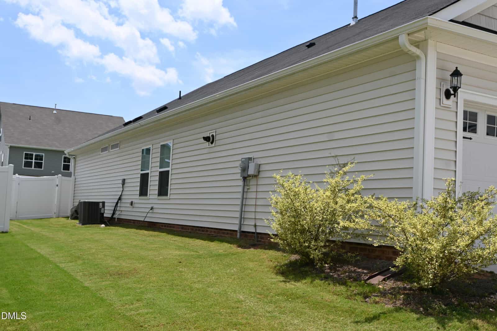 Side view of a white house with horizontal siding, green lawn, some small bushes, air conditioning units, and utility boxes. A white fence and another house are visible in the background under a blue sky.