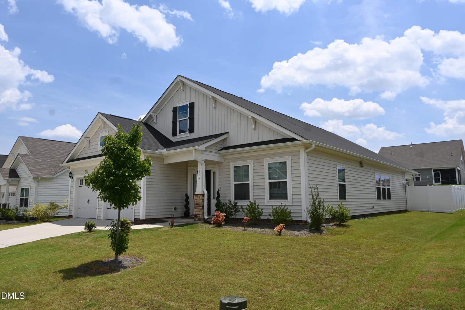 Single-story white house with black shutters, a front porch, attached garage, small tree, and landscaped yard under a blue sky with scattered clouds.