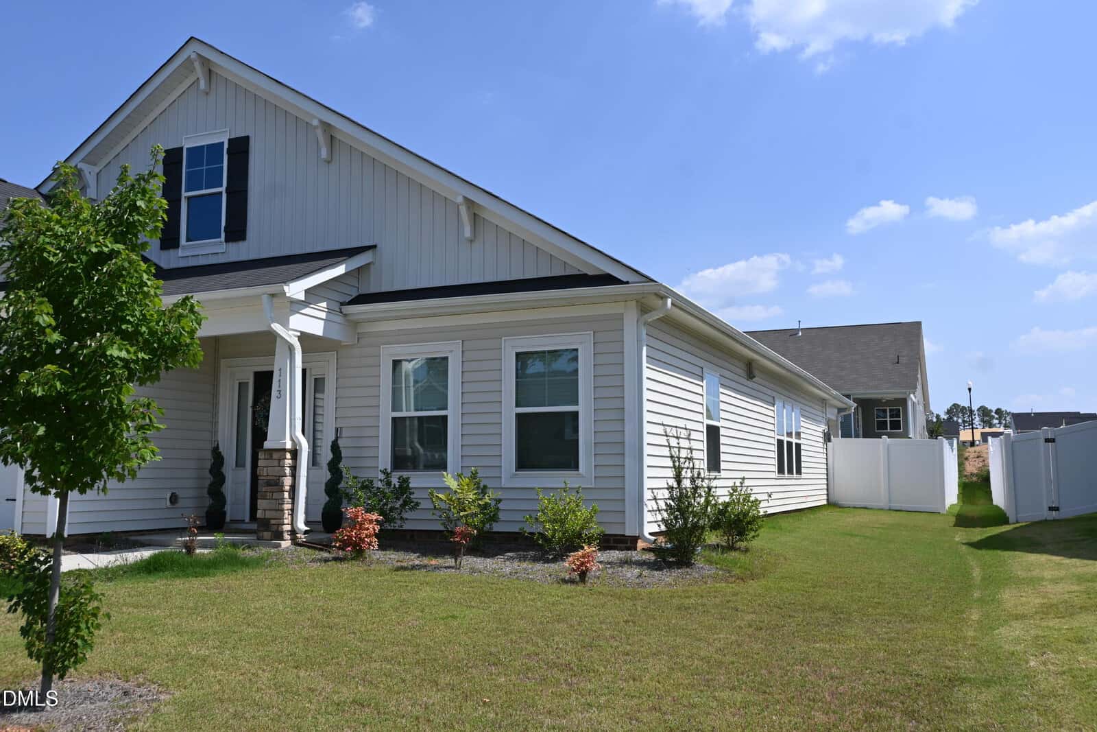 A modern white house with a front porch, green lawn, small shrubs, and a white privacy fence under a clear blue sky.