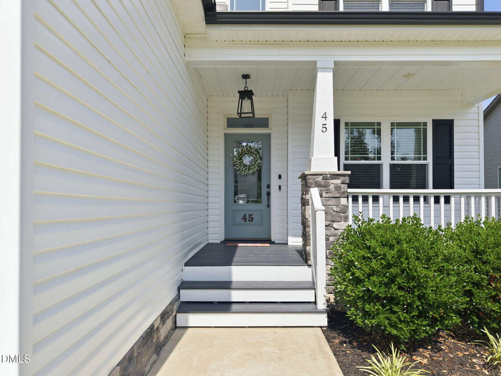 Front porch of a white house with stone accents, a blue door displaying the number 45, a wreath hanging on the door, white railings, and green bushes along the walkway.