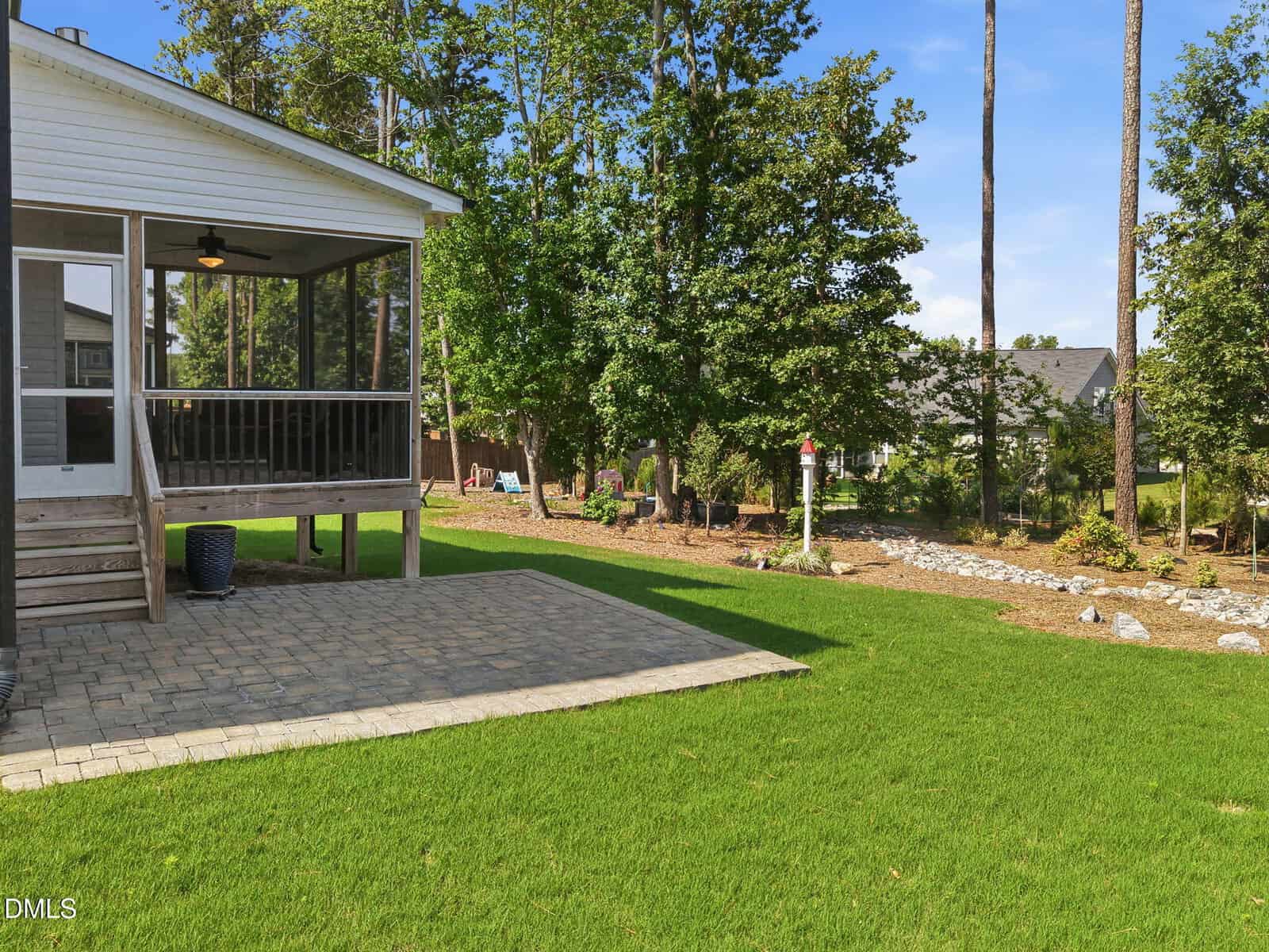 Backyard with a green lawn, stone patio, screened porch, and trees along the fenceline. Rocks and a birdhouse on a post are visible near the back garden area.