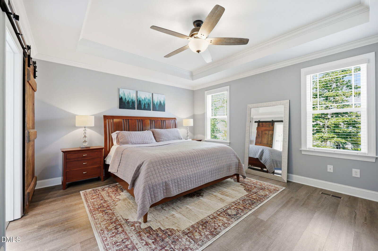 A modern bedroom with a large bed, gray walls, wooden nightstands, a ceiling fan, a floor mirror, artwork above the bed, and two windows letting in natural light.