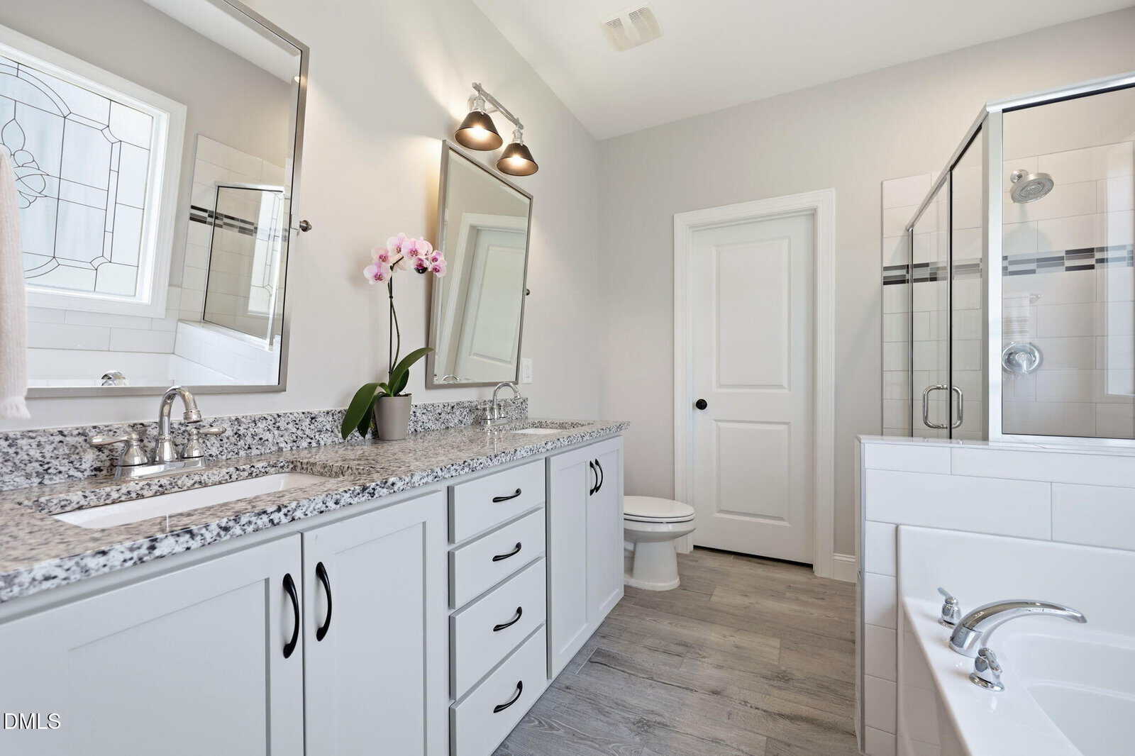 Modern bathroom with double sink vanity, granite countertop, mirrors with lights, an orchid plant, glass shower, bathtub, and white cabinets on light wood flooring. White walls and door complete the clean, bright look.
