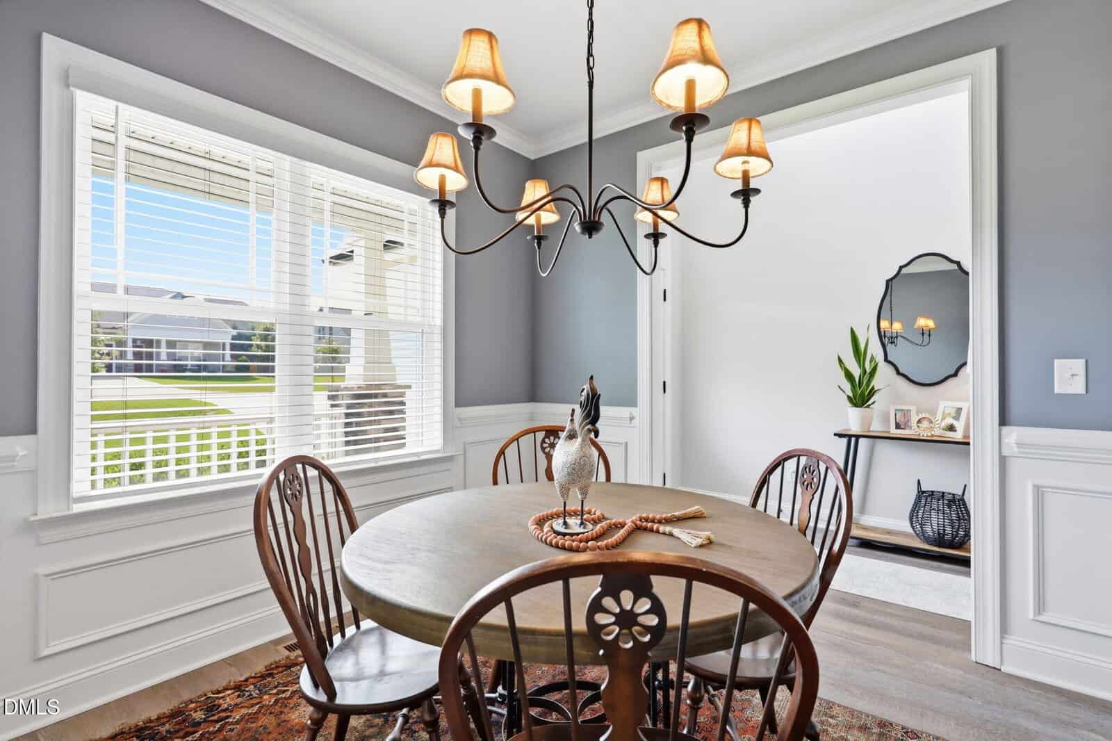 A bright dining room with a round wooden table, four chairs, a rooster centerpiece, and a black chandelier. Light streams in through a large window with blinds, and an entryway with decor is visible in the background.