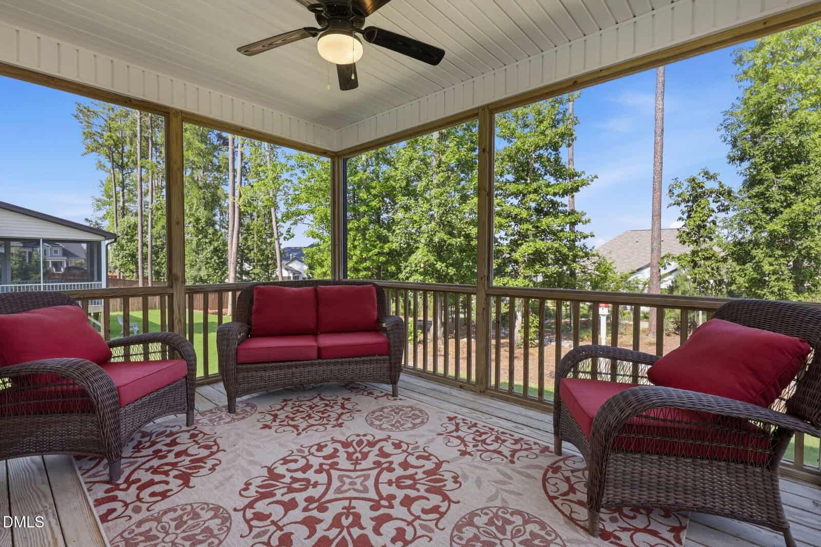 Screened-in porch with wicker furniture featuring red cushions and a patterned area rug. Ceiling fan overhead. Large windows show a green backyard with trees and neighboring houses visible in the background.