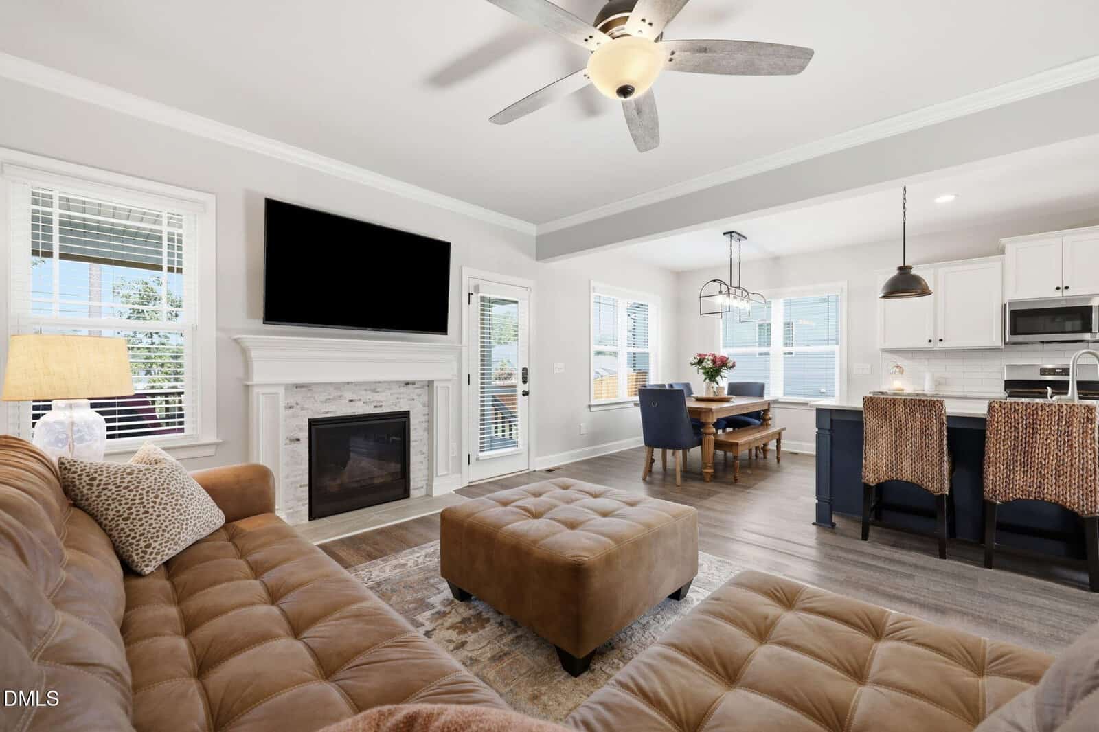 A modern living room with tan sofas, a fireplace with a TV above it, and an ottoman. Large windows let in natural light. The open kitchen features a dining table with black chairs and pendant lights above an island.