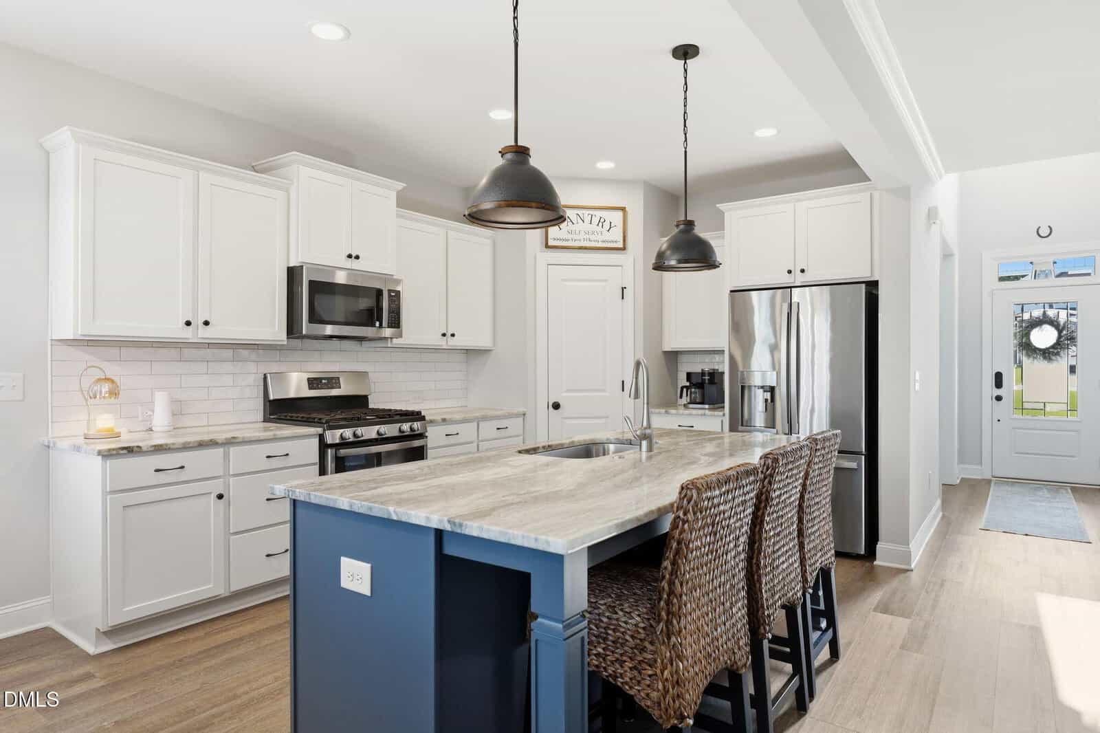 Modern kitchen with white cabinets, stainless steel appliances, a blue island with a marble countertop, three woven barstools, pendant lighting, wood floors, and a door leading to a hallway with a windowed front door.