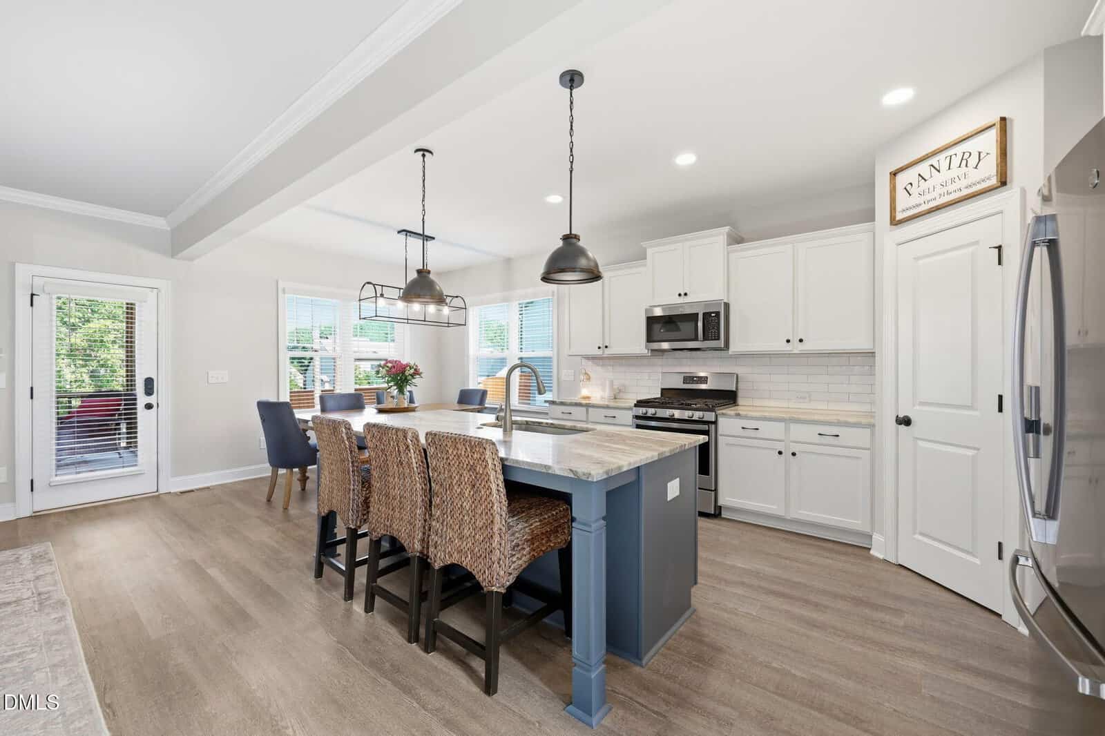 Modern kitchen with an island featuring a sink, three wicker barstools, stainless steel appliances, white cabinets, pendant lighting, a dining table with blue chairs, and a door leading to an outdoor patio.