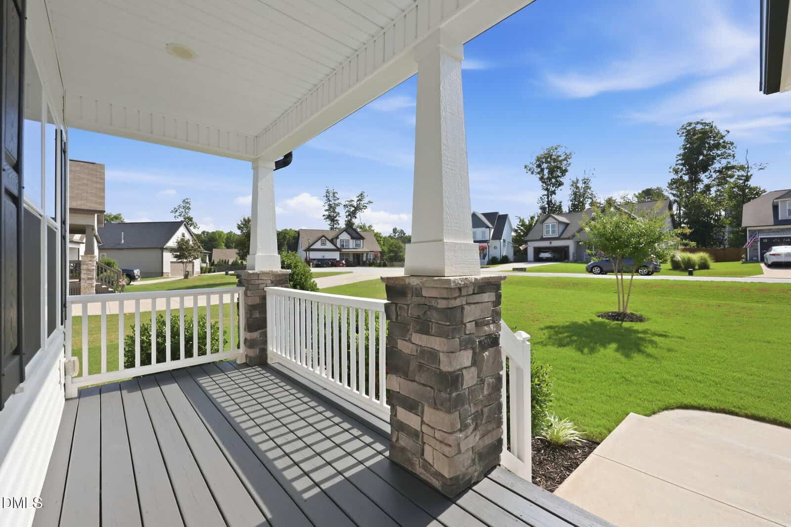 View from a covered front porch with white railings and stone columns, overlooking a neatly landscaped suburban neighborhood with green lawns, trees, and houses under a blue sky.