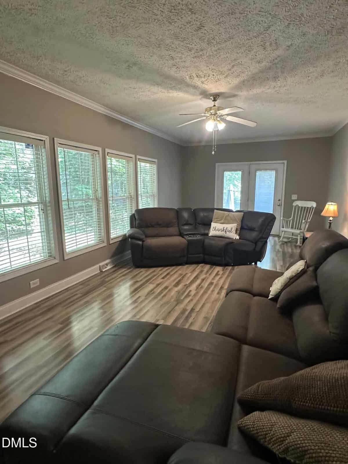 A living room with brown sofas, a “Thankful” pillow, wood flooring, large windows, a ceiling fan with light, and a small white table with chairs near glass doors leading outside.