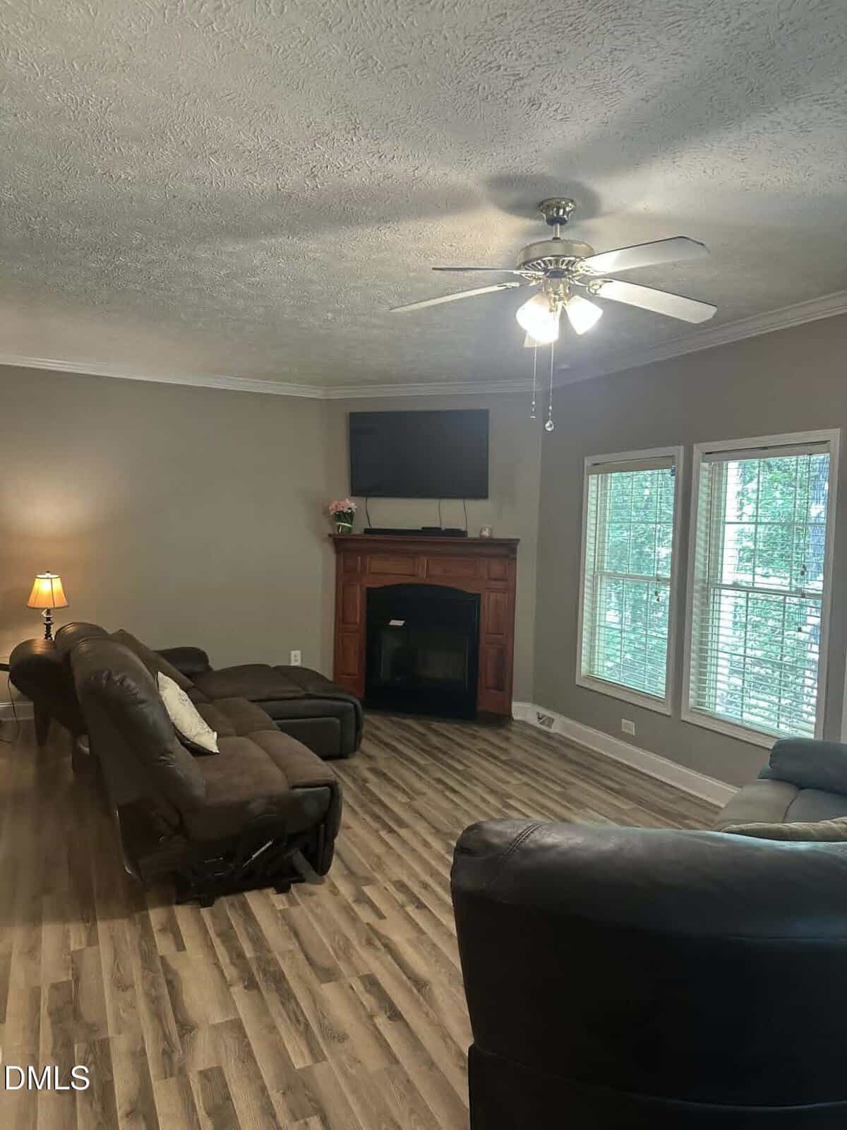 A living room with brown recliners, wood flooring, a ceiling fan with lights, a fireplace with a TV above it, large windows, and a small lamp on a side table.