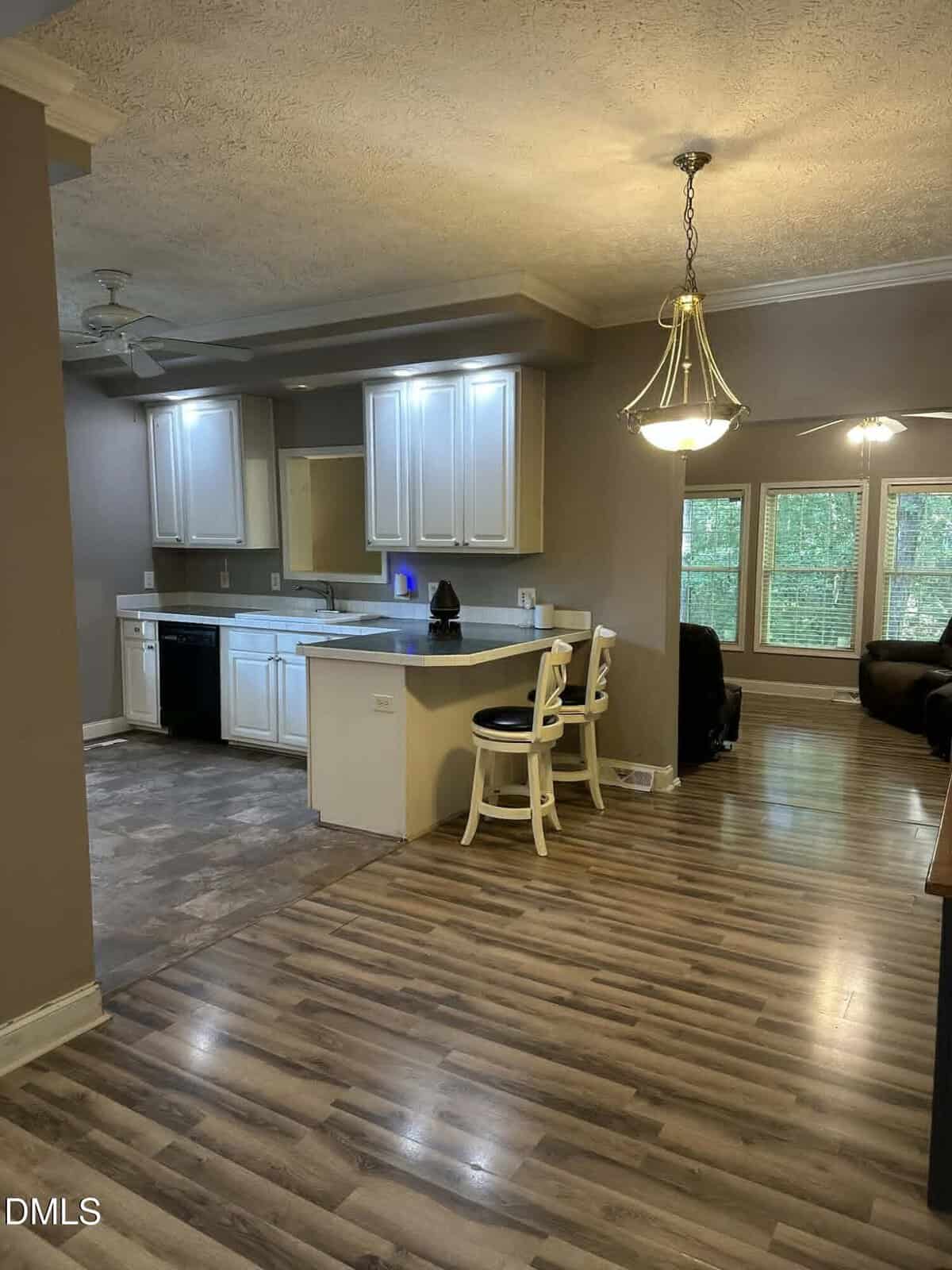 Open kitchen with white cabinets, two white barstools at a counter, and a hanging light fixture. Adjacent living area has large windows with views of trees. Wood and tile flooring throughout.