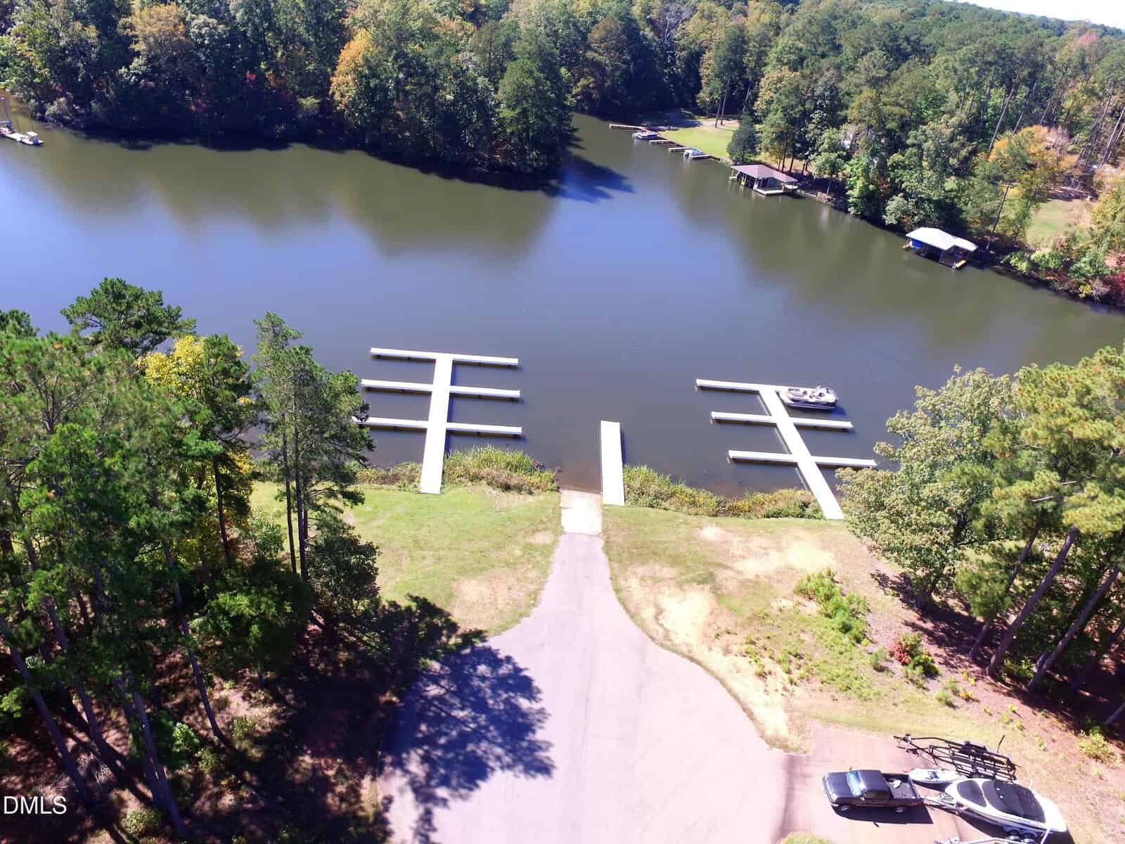 Aerial view of a boat launch and multiple empty docks on a calm, tree-lined lake, with a road leading to the water and a parked car and trailer nearby.