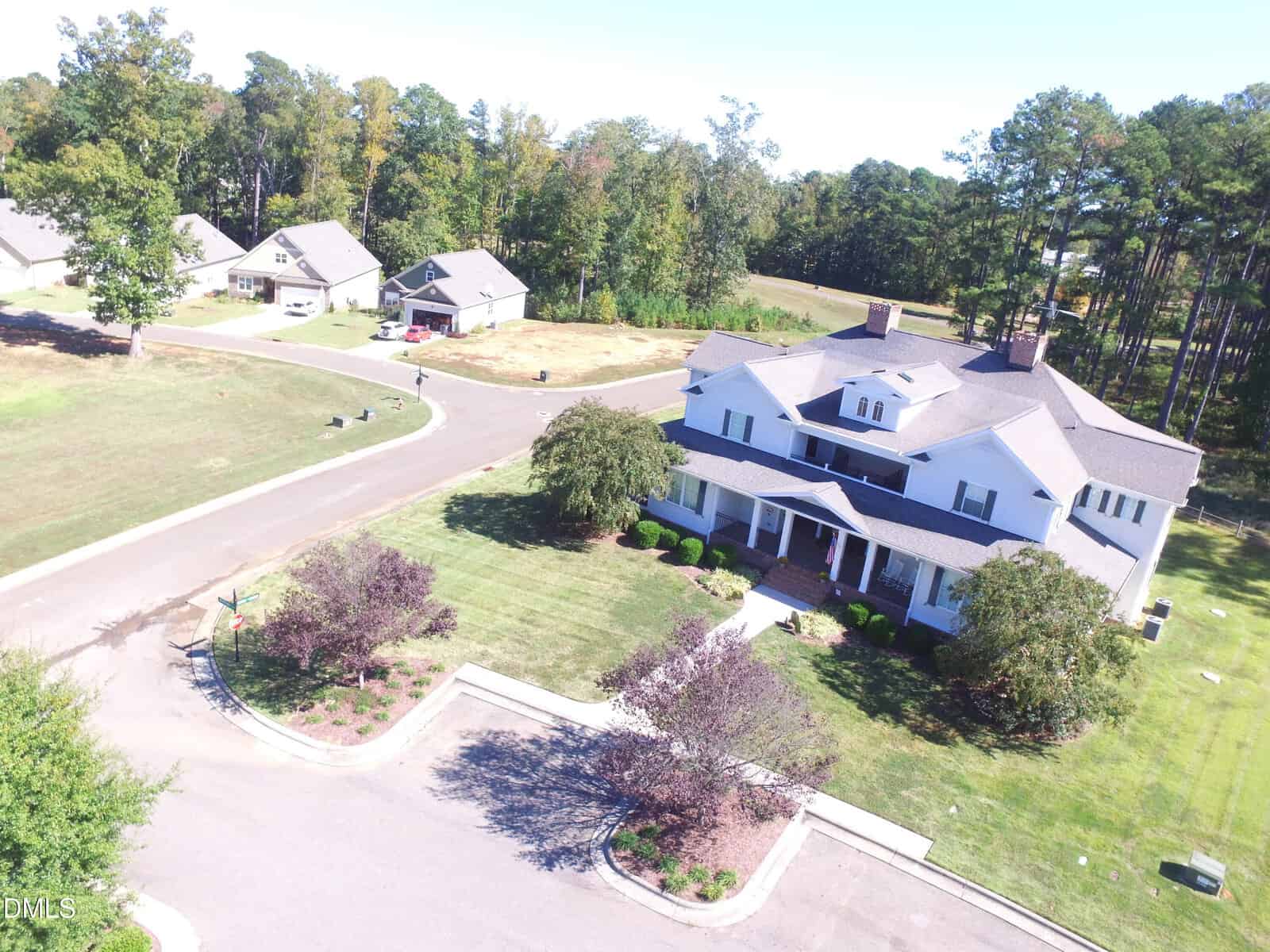 Aerial view of a large white house with a front porch and manicured lawn, situated on a corner lot in a suburban neighborhood with nearby homes and trees.
