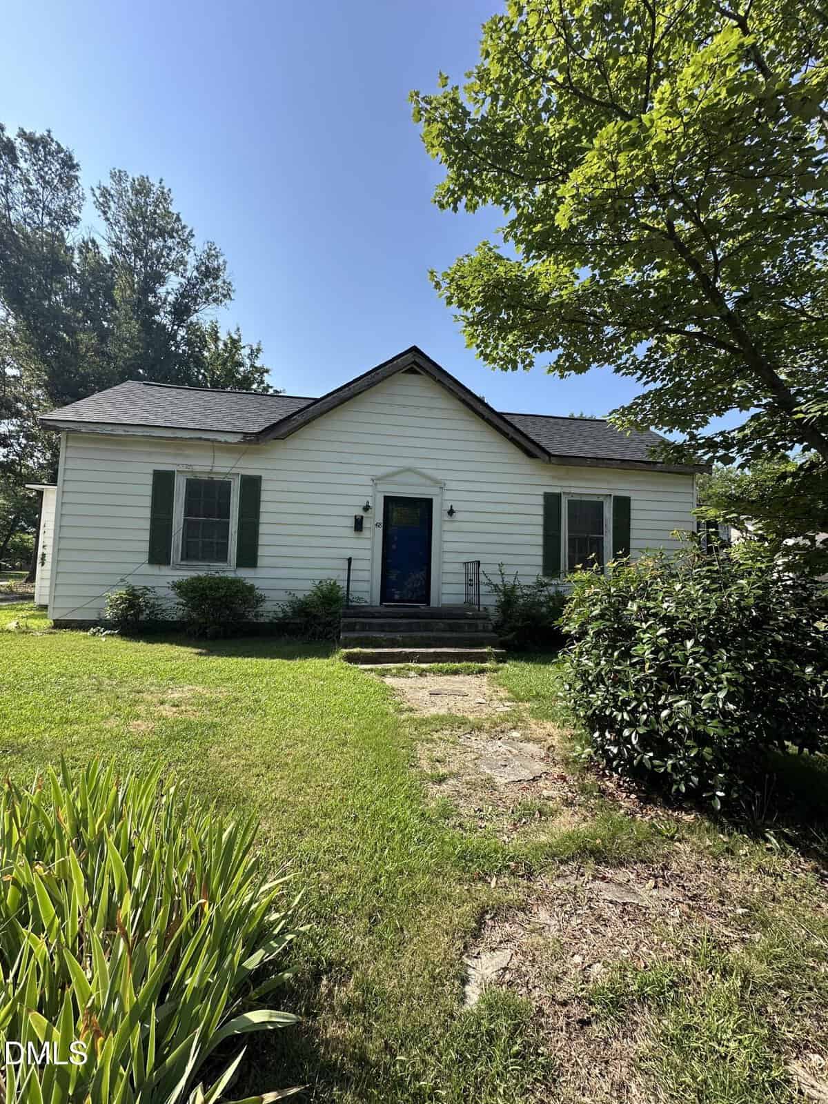 A small white house with green shutters, a blue front door, and a gabled roof sits behind a green lawn and shrubs, with a tree partially shading the right side of the yard on a sunny day.