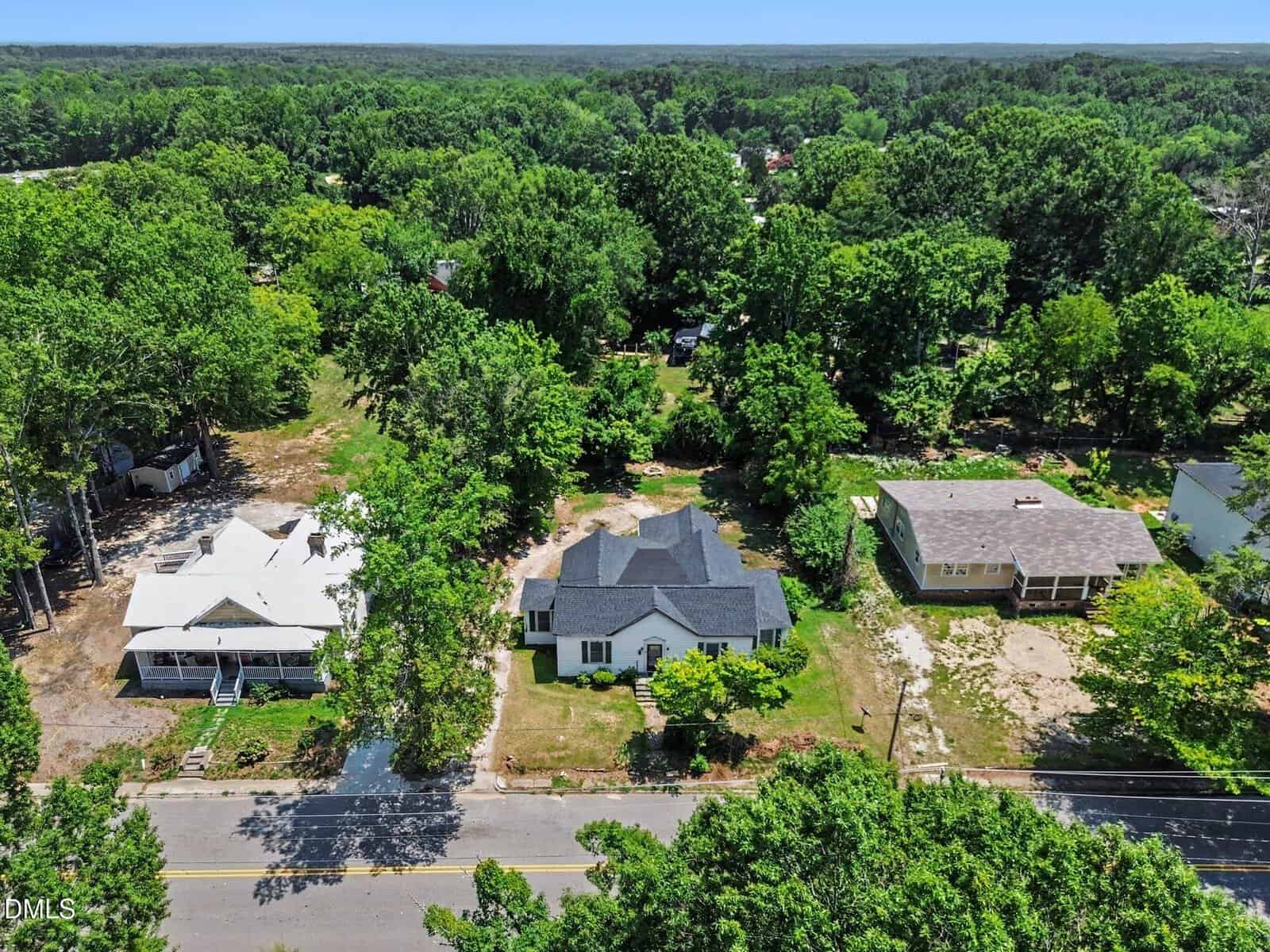 Aerial view of three houses along a tree-lined street, surrounded by lush greenery and dense forest in the background on a sunny day.