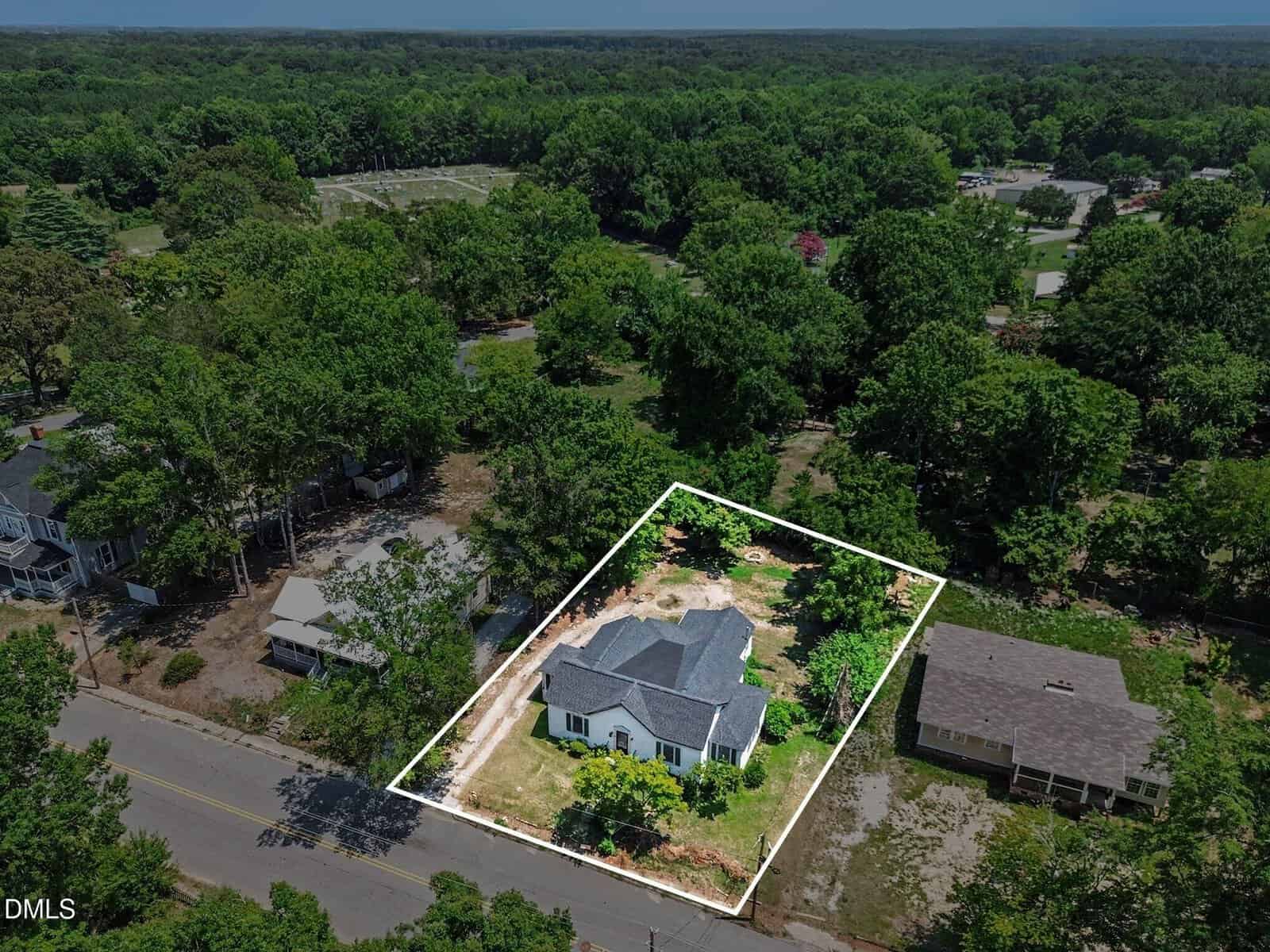 Aerial view of a single-story white house with a dark roof, outlined by a white border, surrounded by trees and neighboring homes in a wooded residential area.