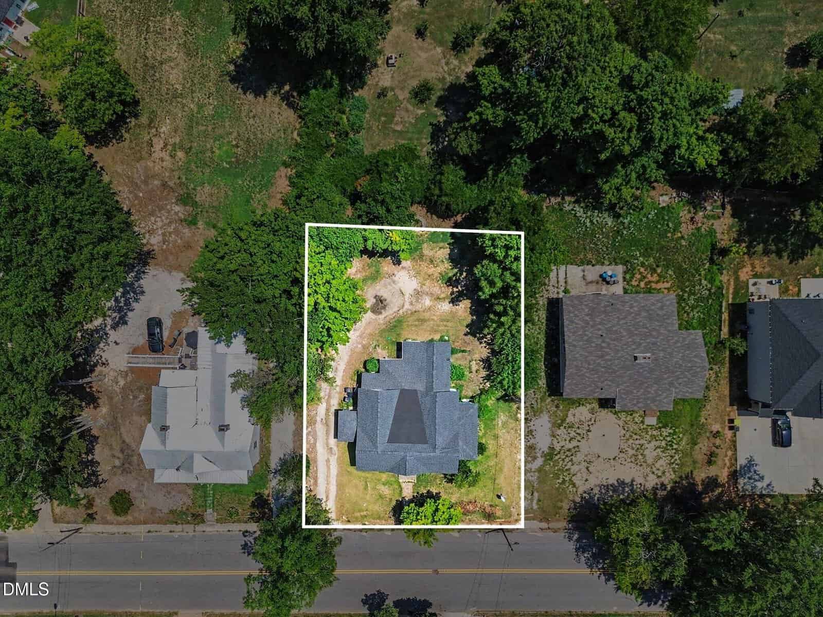 Aerial view of a house with a gray roof, centered on a rectangular lot outlined in white, surrounded by trees and neighboring homes on either side. A dirt driveway leads from the street to the house.