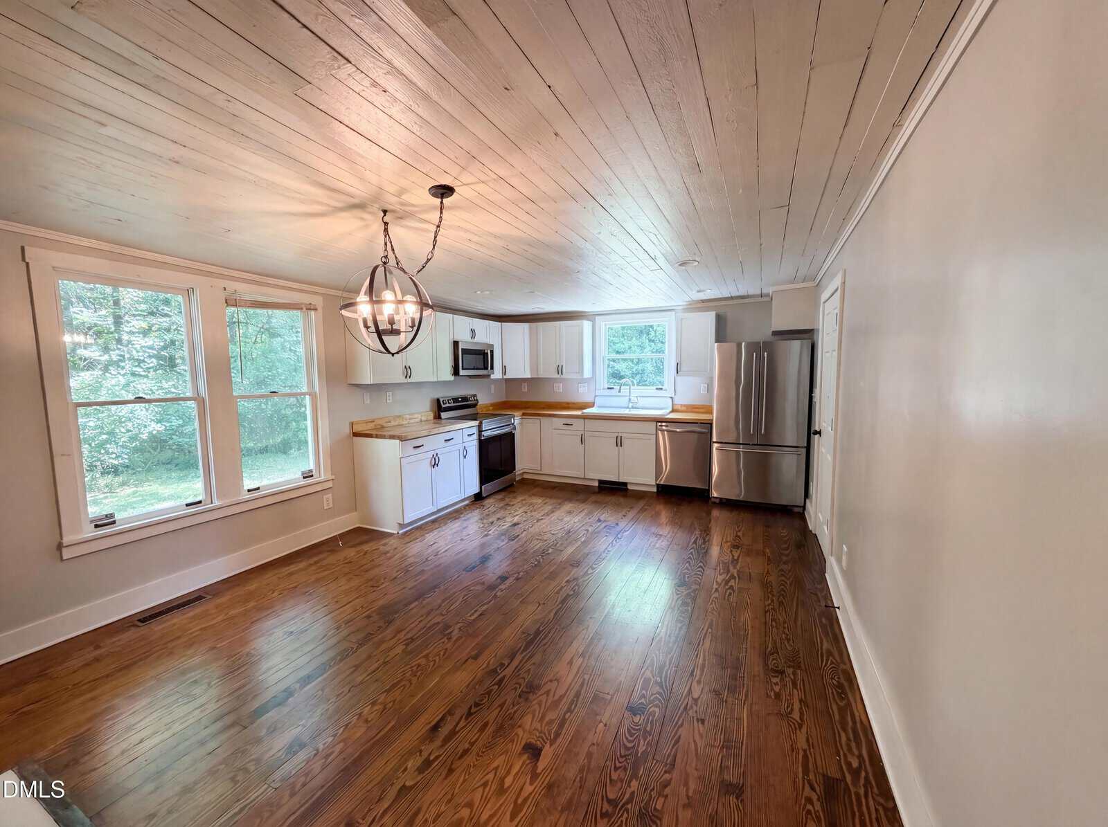 Bright, open kitchen and dining area with wood floors, white cabinets, stainless steel appliances, large windows, and a modern chandelier. The ceiling is white with a rustic, wooden texture.