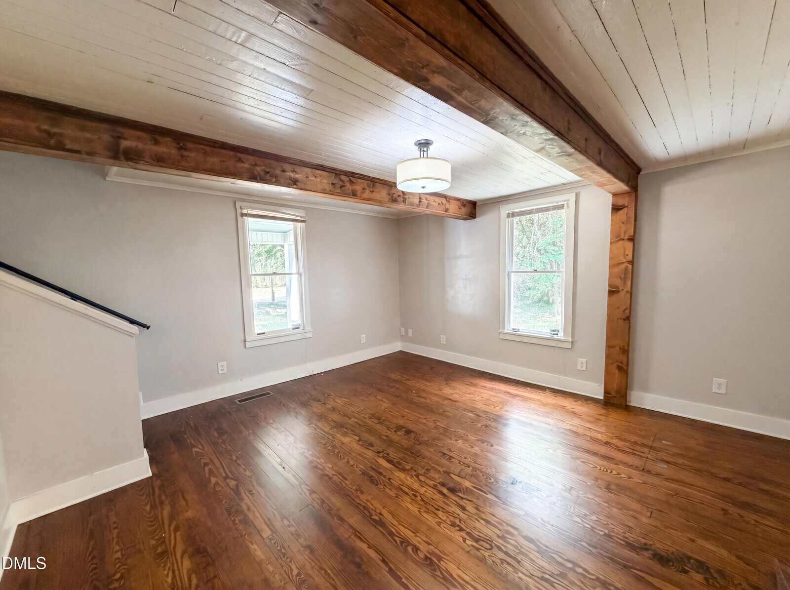 A bright, empty room with wooden floors, white walls, exposed wood ceiling beams, two large windows, and a modern ceiling light fixture. Natural light streams in through the windows.