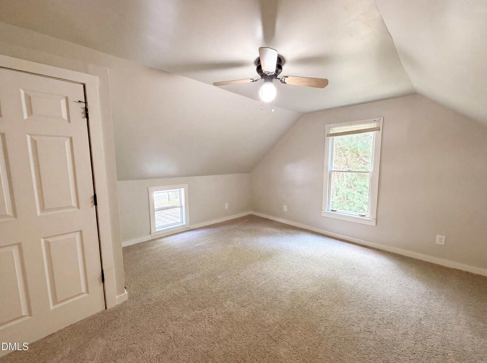 A bright attic bedroom with sloped ceilings, light beige walls, and carpeted floor. The room has a ceiling fan with a light, a white door, one large window, and a smaller window near the floor.
