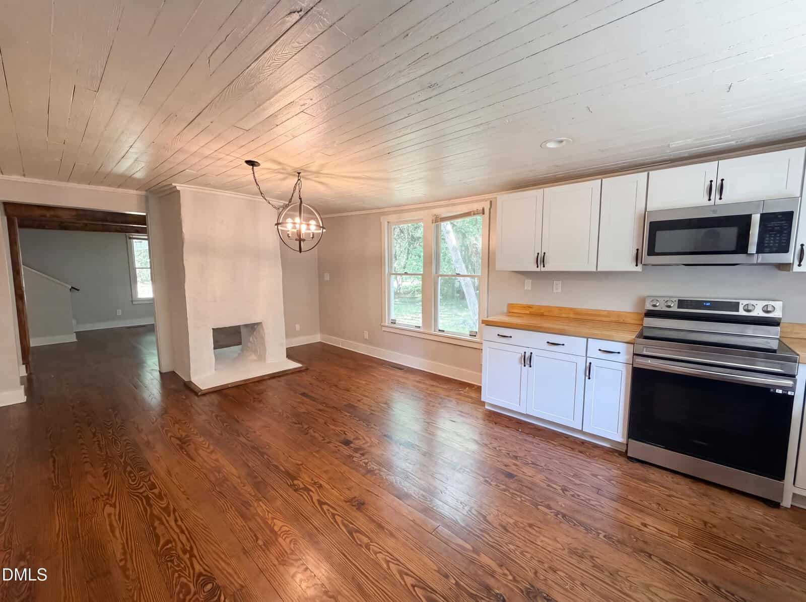 Bright kitchen with white cabinets, stainless steel appliances, wood countertops, chandelier, wood floors, large window, and a white painted brick fireplace in an open living space.
