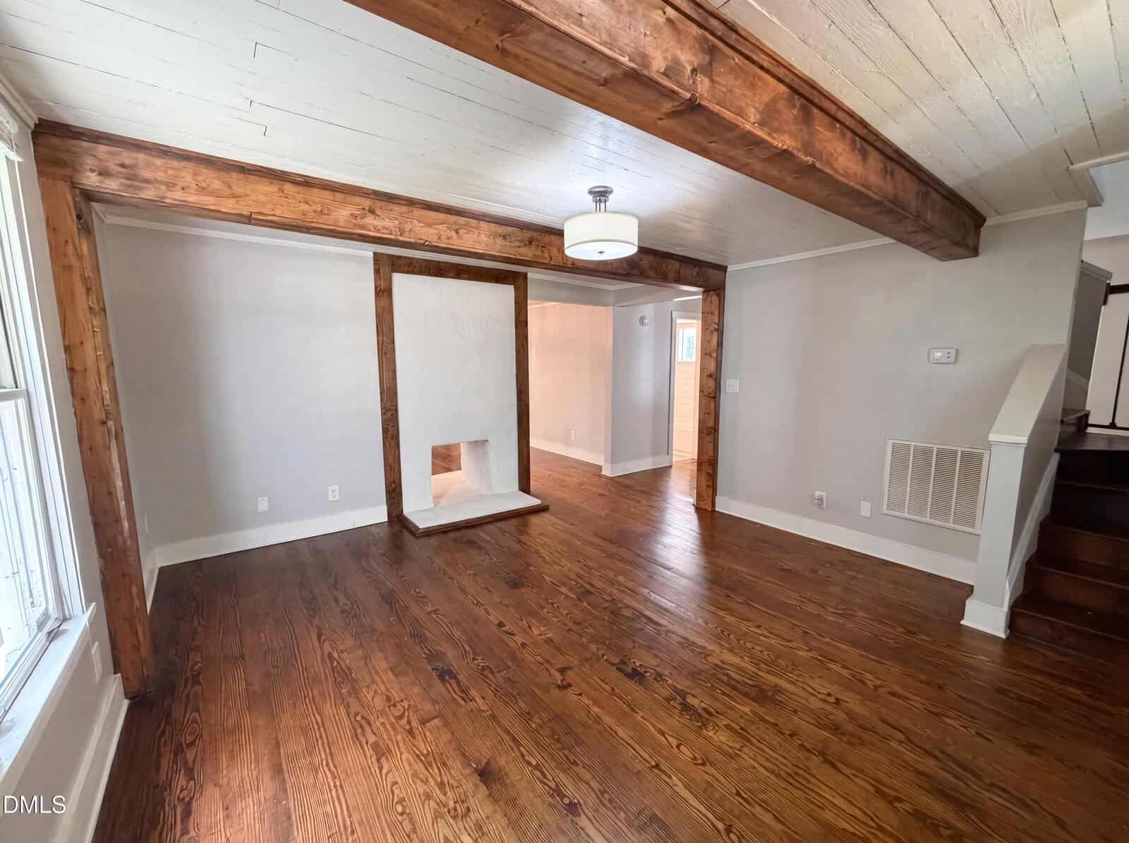 A spacious living room with wood floors, exposed wood beams, white walls, a ceiling light, and a unique framed wall opening. Stairs are visible to the right.