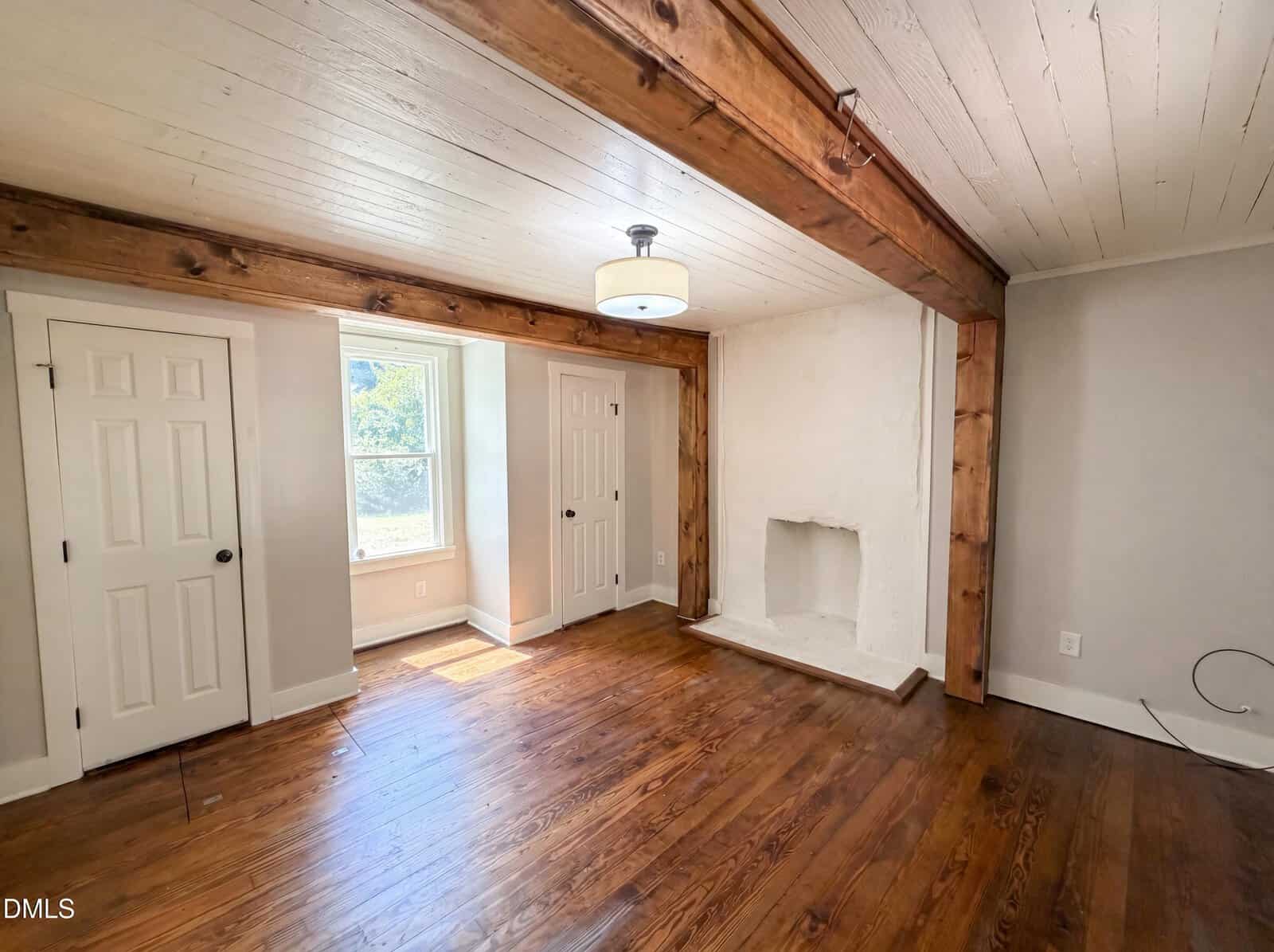 Bright room with hardwood floors, wood ceiling beams, two white doors, a window letting in sunlight, light fixture, and a white painted fireplace on the right wall. Walls are light gray and the ceiling is white.