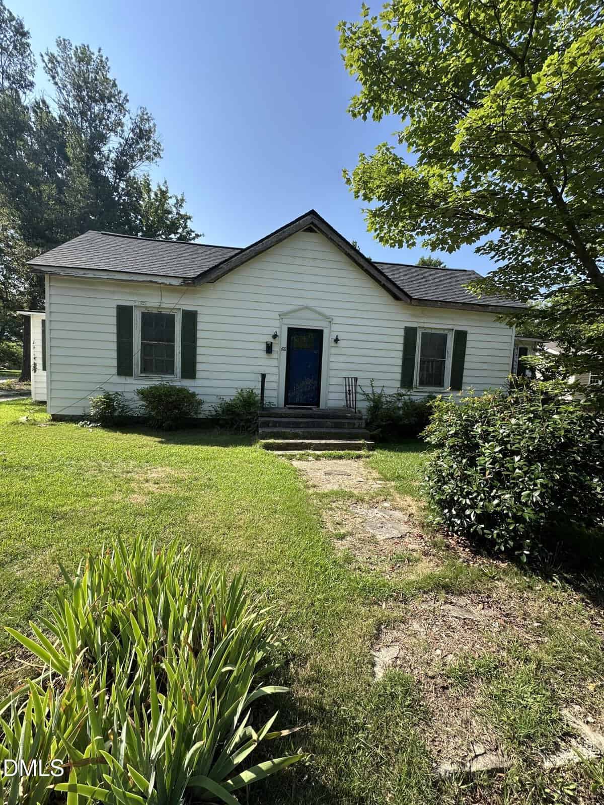 A small white house with green shutters and a blue front door sits on a grassy lawn at 418 E Green Street, Franklinton, surrounded by shrubs and trees under a clear blue sky. Concrete steps lead up to the entrance.