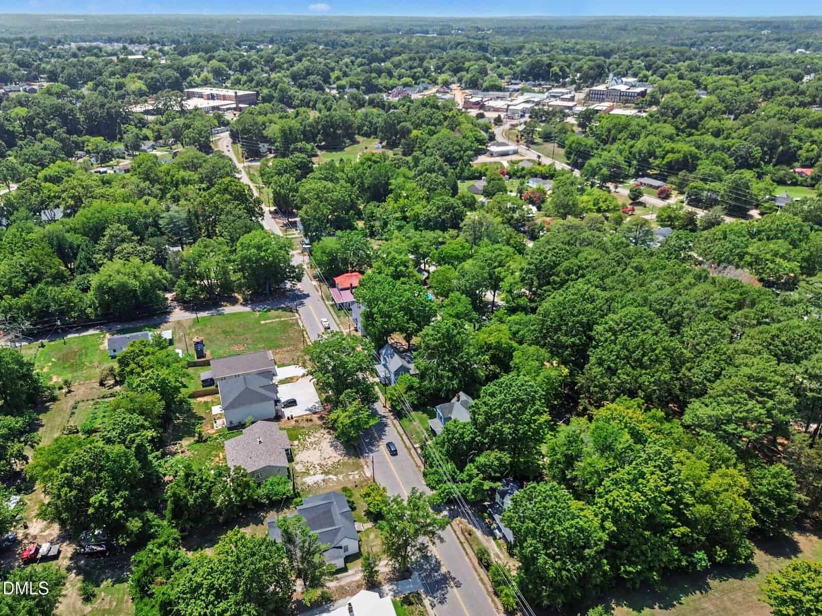 Aerial view of a suburban neighborhood with houses, roads, and dense green trees on a sunny day, extending toward a distant horizon.