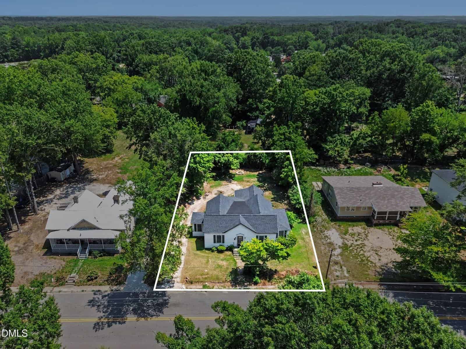 Aerial view of a white house outlined in white, centered on a grassy lot with trees surrounding it. Two neighboring homes sit on either side, and a road runs across the bottom of the image. Dense forest fills the background.