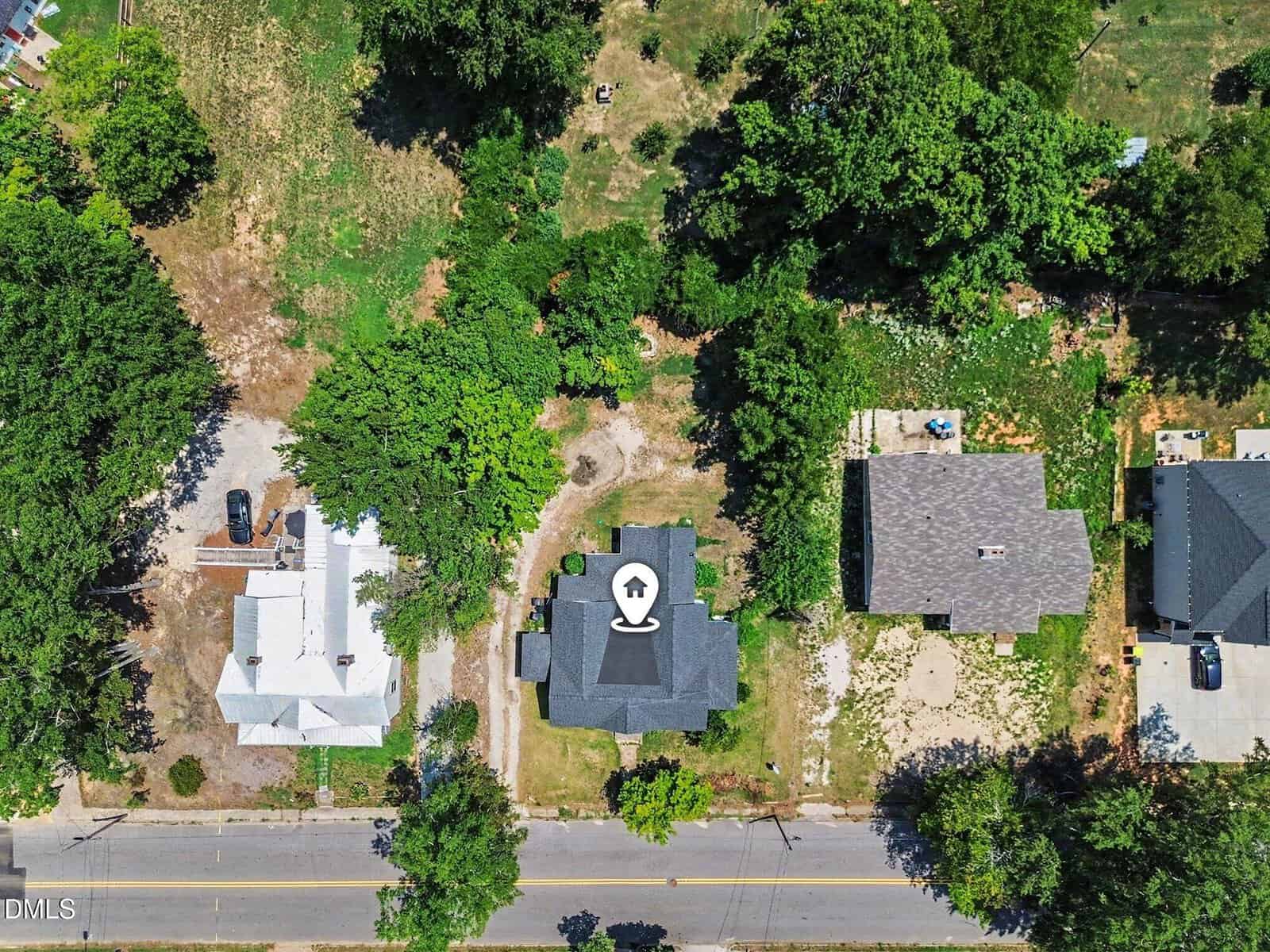 Aerial view of three houses along a road, with the middle house highlighted by a location marker icon on its roof. Surrounding areas have trees, grass, and driveways.