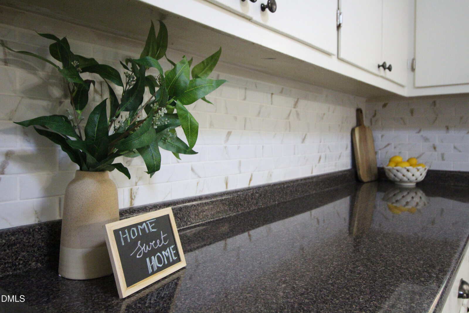 A kitchen counter with a potted plant, a small “Home Sweet Home” chalkboard sign, a cutting board, and a bowl of lemons against a white tiled backsplash.