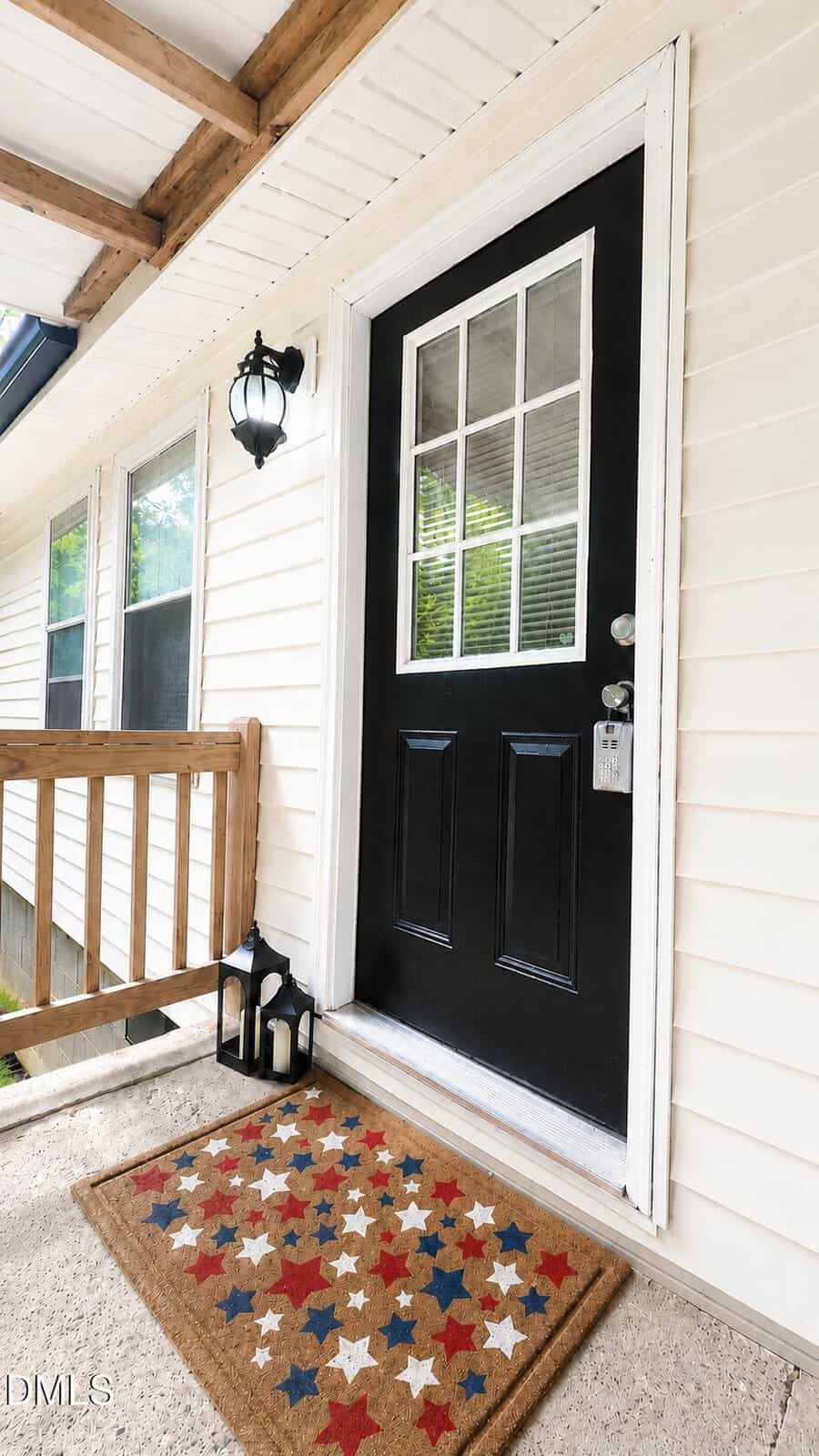 A black front door with glass panes, a keypad lock, and a welcome mat decorated with red, white, and blue stars. Two lanterns sit beside the door on a tan porch of a light-colored house.