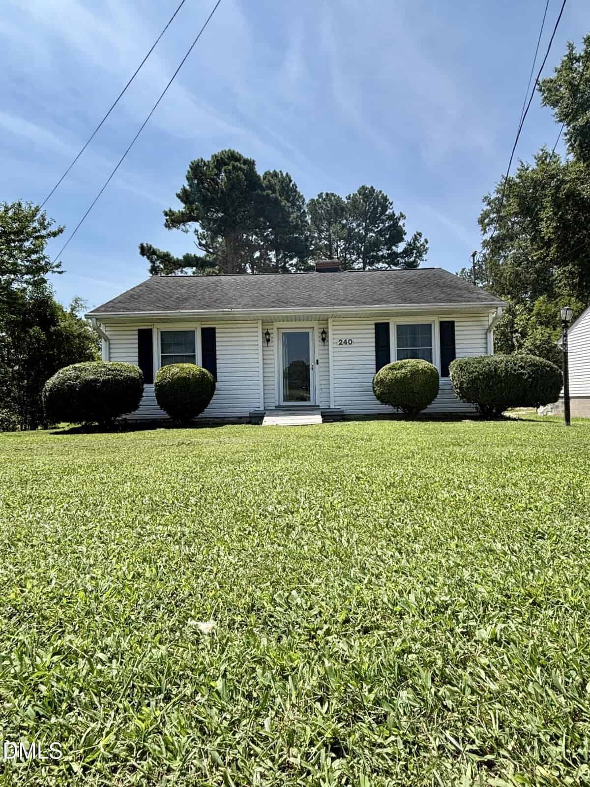 Small white single-story house with dark shutters, black roof, central front door, and trimmed bushes. Located at 240 Vicksboro Road in Henderson, this home sits on a well-maintained green lawn with trees in the background and a clear sky above.