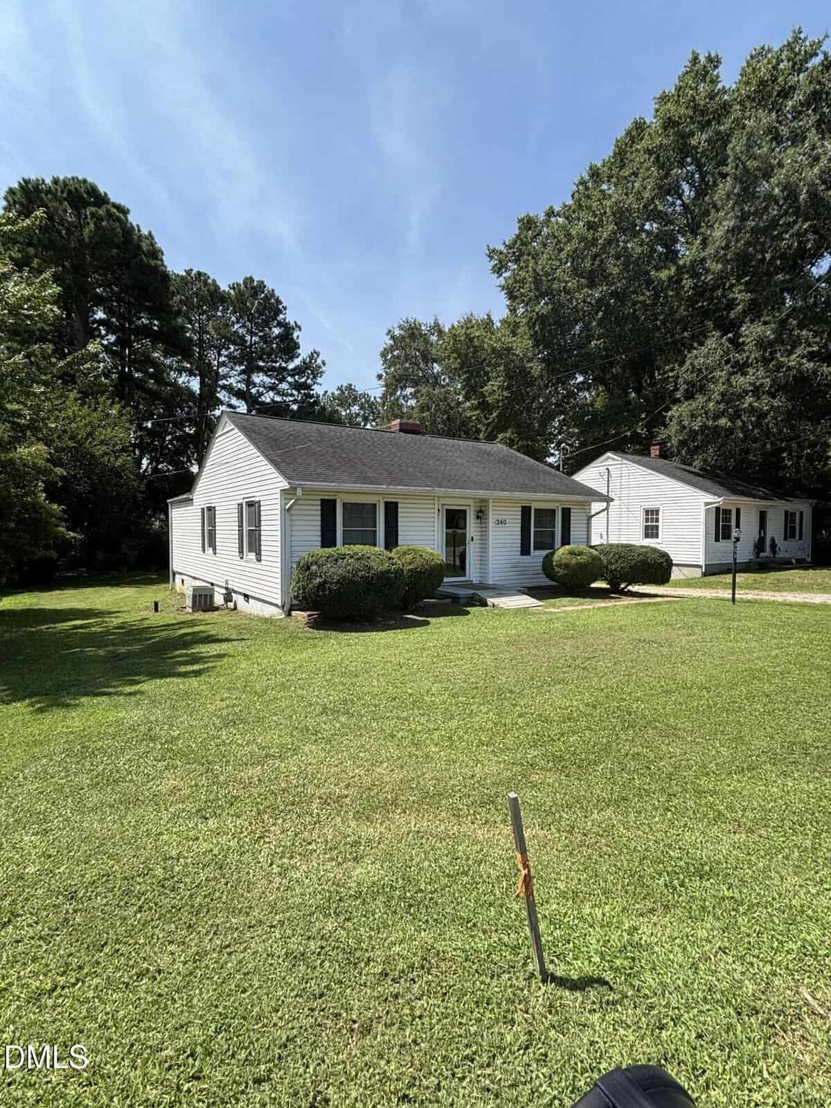 Single-story white house with black shutters and a small front porch, surrounded by neatly trimmed bushes and a large, grassy yard. Another similar house is visible to the right, with trees in the background under a clear sky.