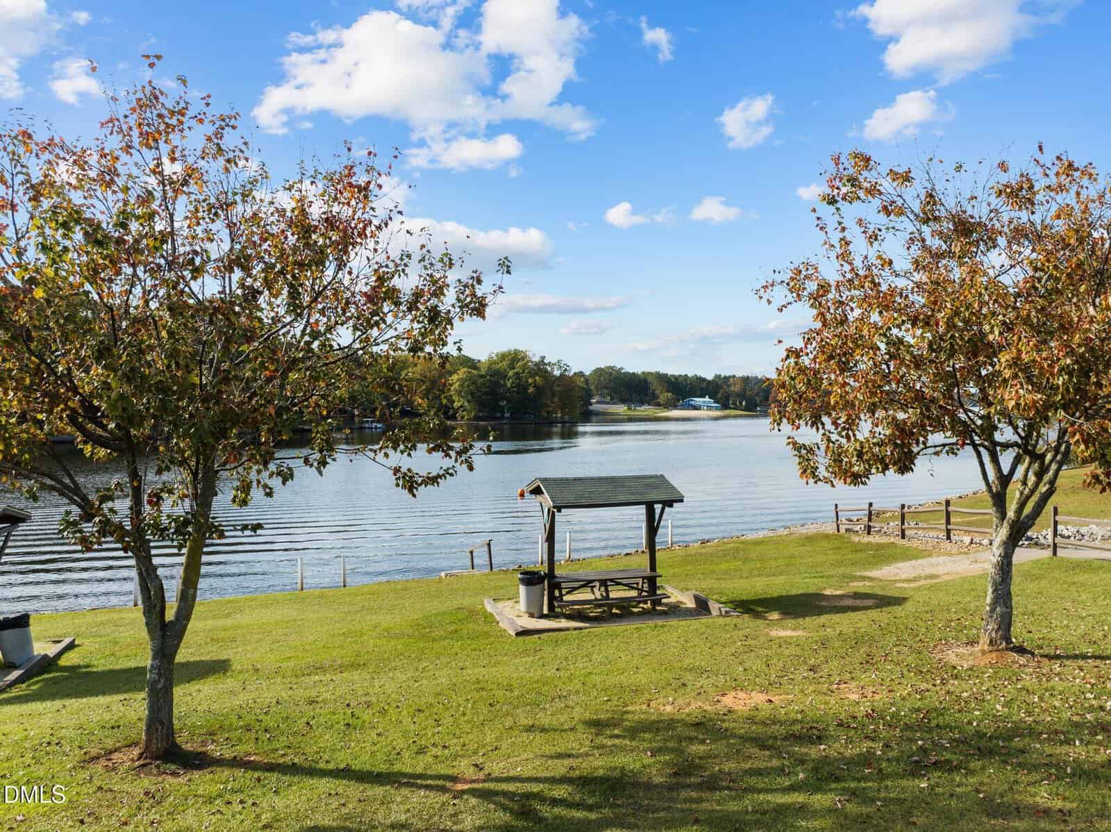A wooden picnic table with a roof sits between two trees on a grassy area overlooking a calm lake under a blue sky with scattered clouds. A fence runs along the lake’s edge.