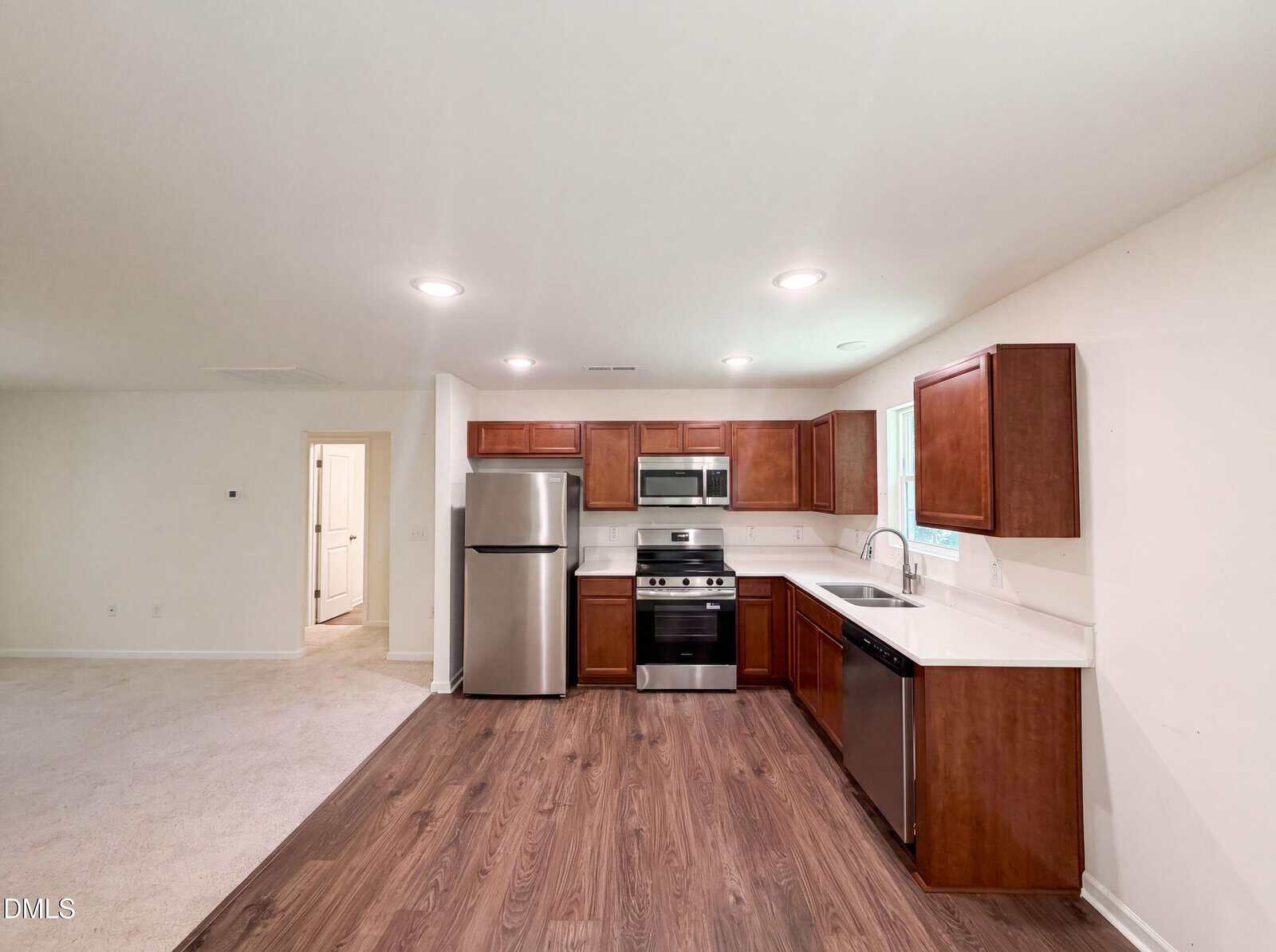 Modern kitchen with stainless steel appliances, wooden cabinets, white countertops, and wood-look flooring, opening into a bright carpeted living area with doorways leading to other rooms.