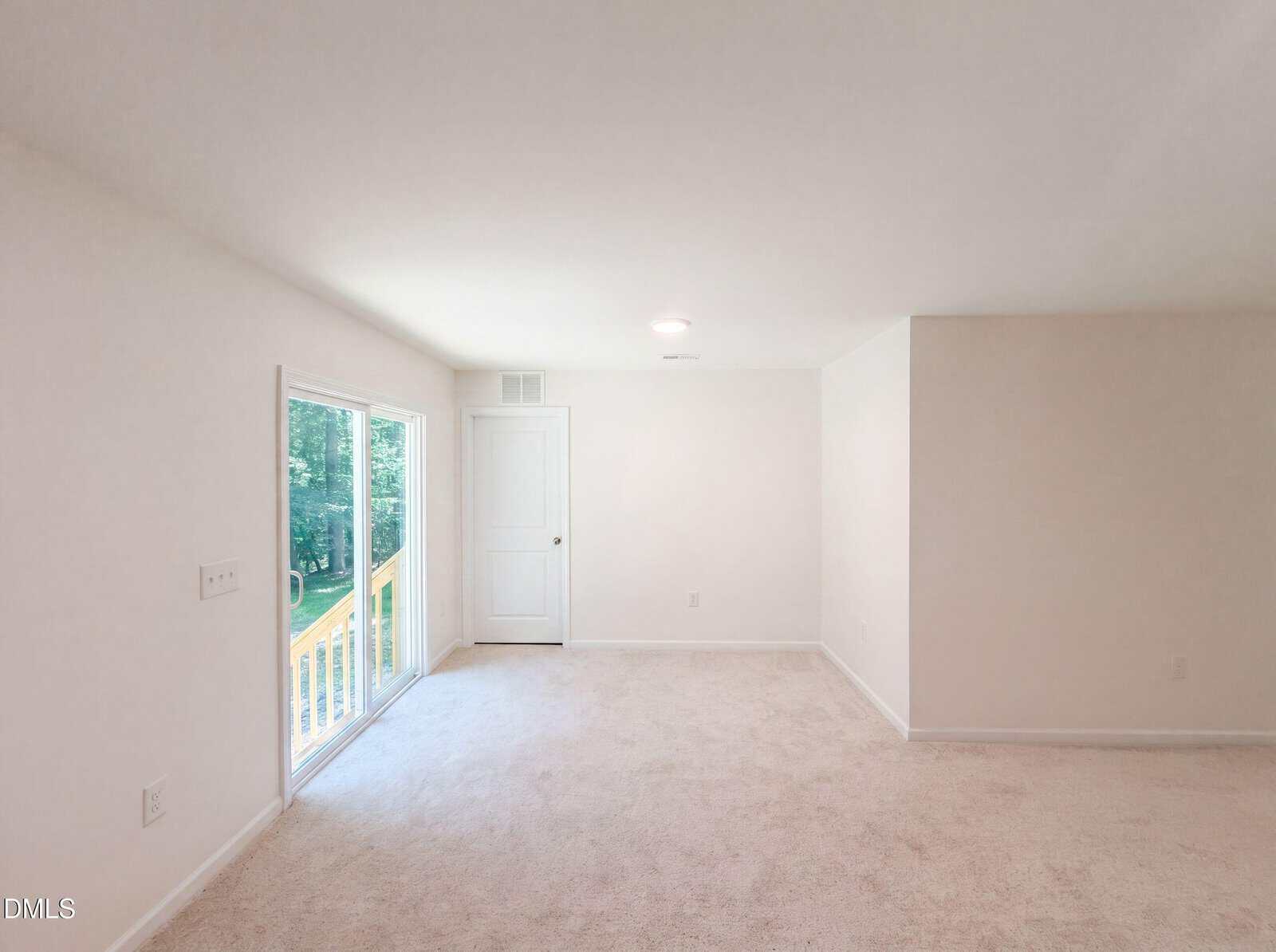A bright, empty room with light beige carpet, white walls, a closed white door, and large sliding glass doors opening to a wooden balcony with a view of trees outside.